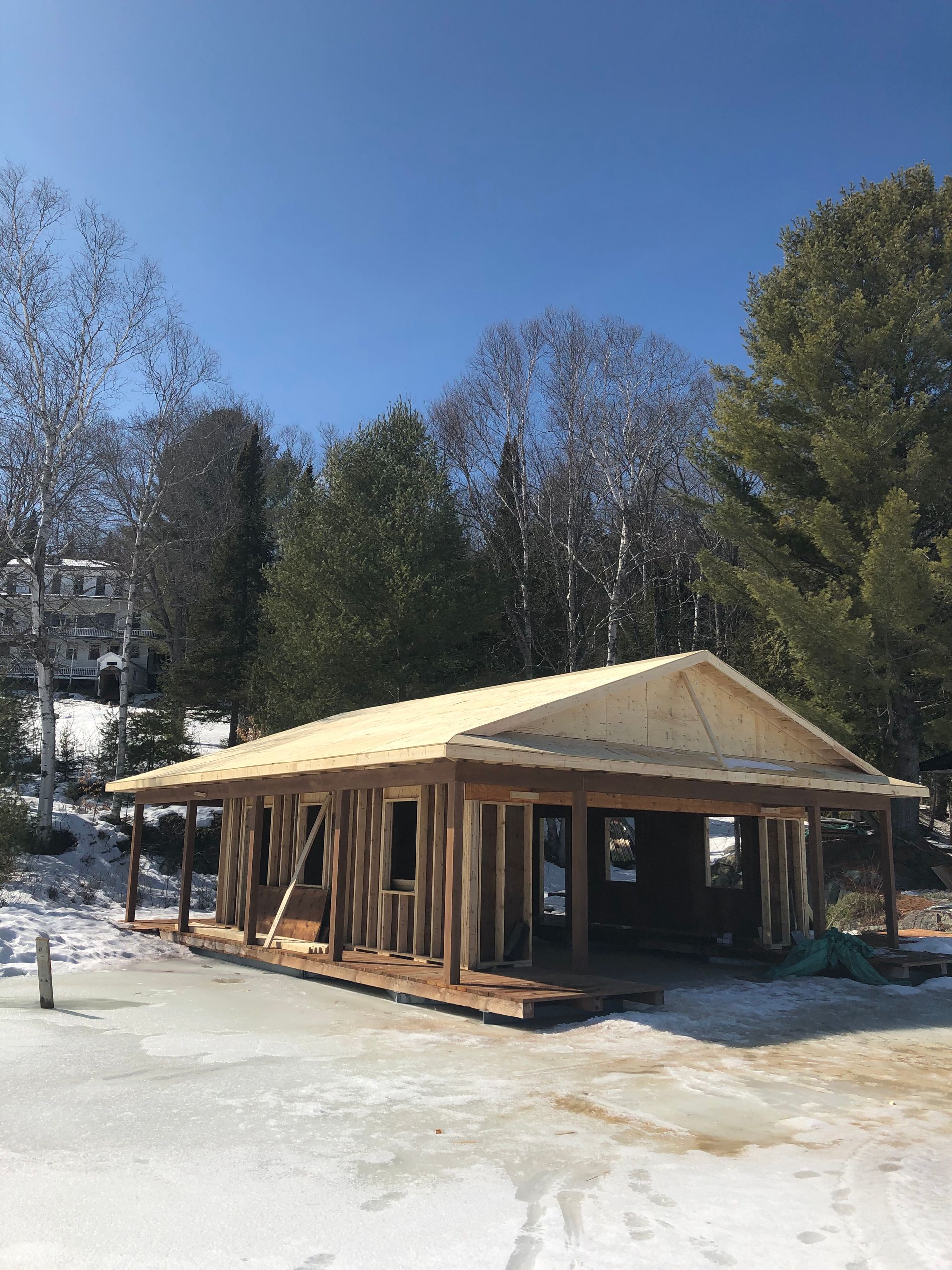 Une cabane en bois en construction dans une clairière enneigée, entourée de grands arbres, sous un ciel d'un bleu éclatant.