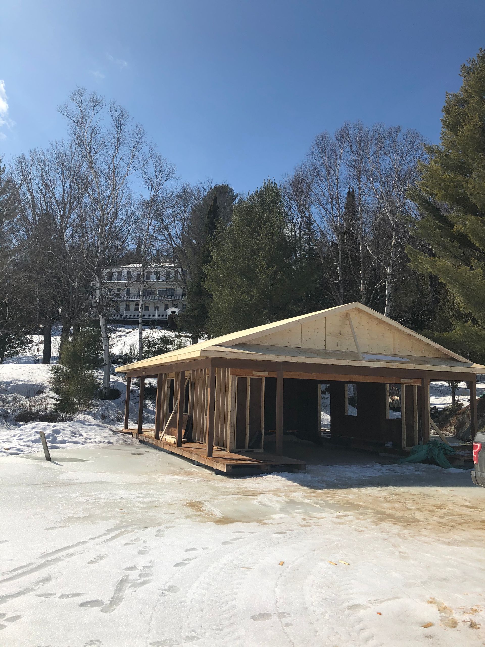 Une structure en bois en construction se dresse sur un sol enneigé, devant des arbres et une maison sur une colline.