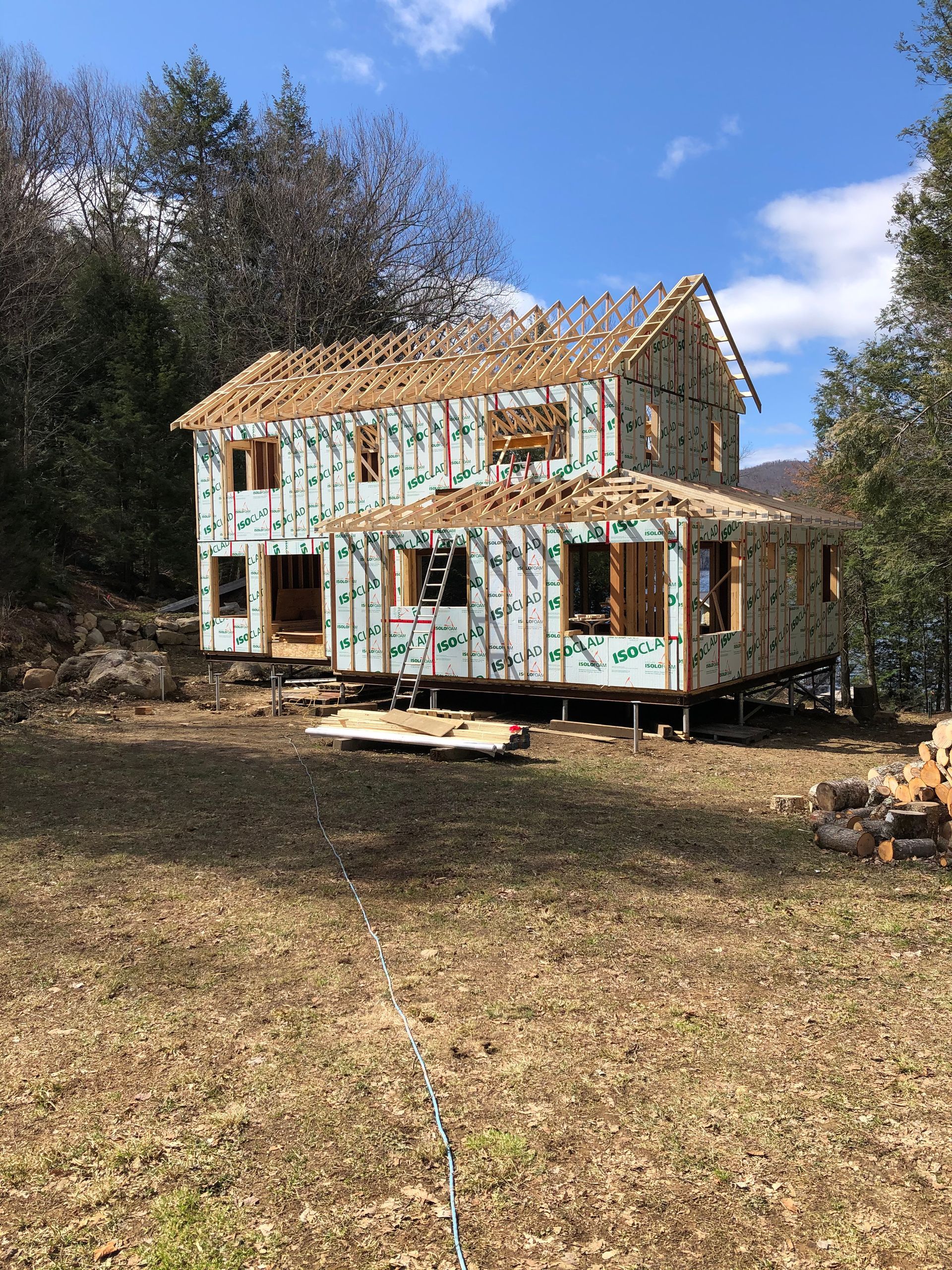 Une maison en construction, avec une charpente en bois apparente et des planches de revêtement, sur un terrain ensoleillé.