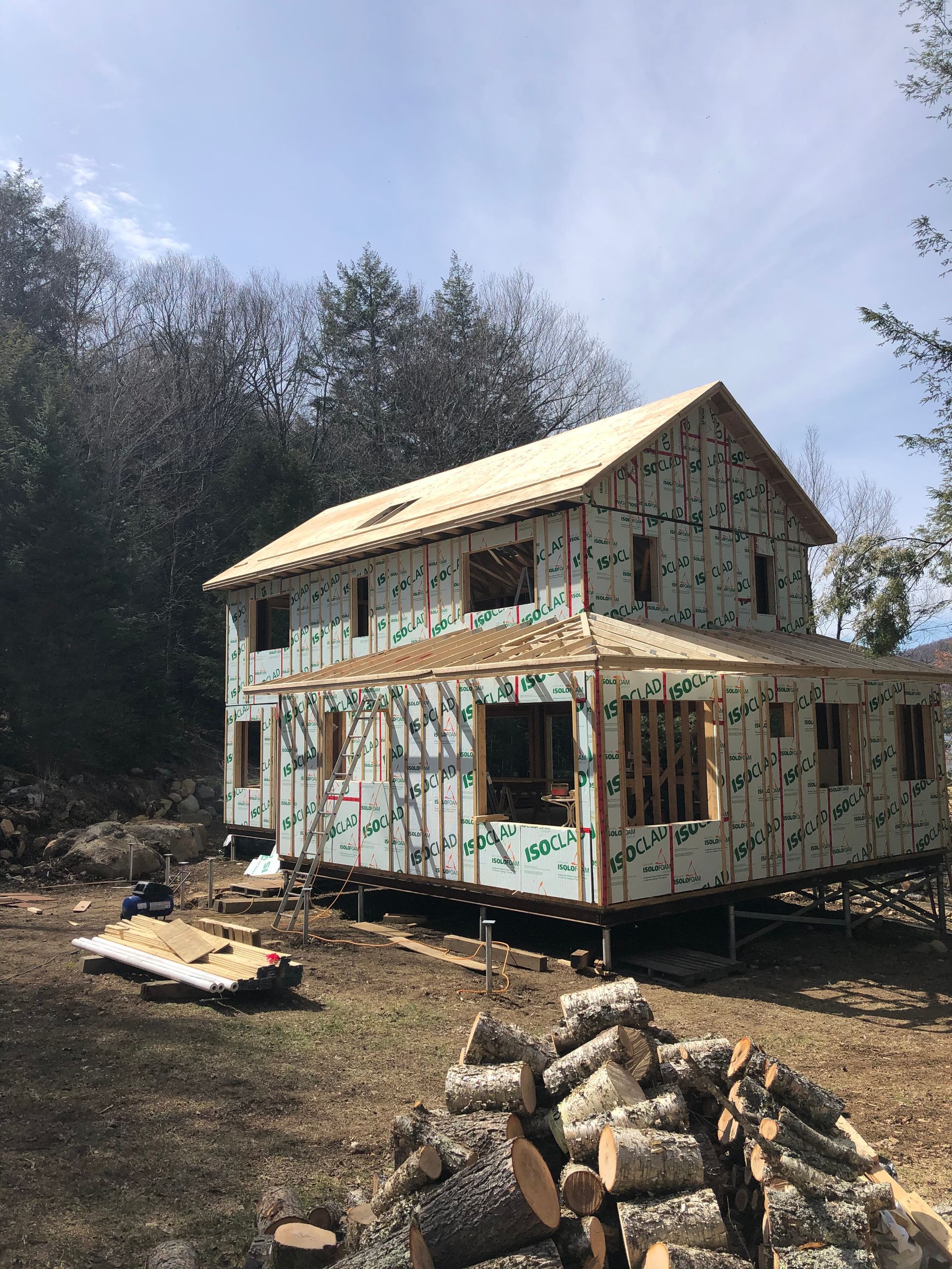 Une maison en construction, recouverte de panneaux isolants blancs, avec un toit en bois et des bûches empilées dans la cour.