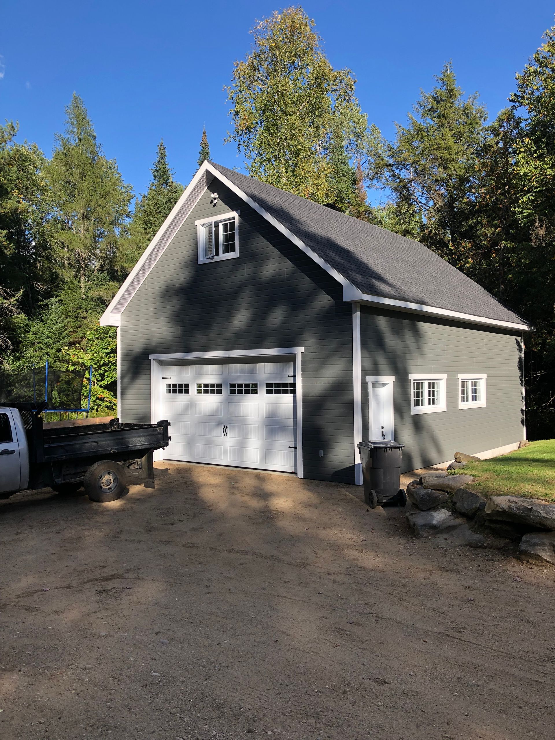 Un garage gris à deux étages avec une porte et des boiseries blanches, entouré d'arbres sur un terrain de gravier sous un ciel bleu azur.