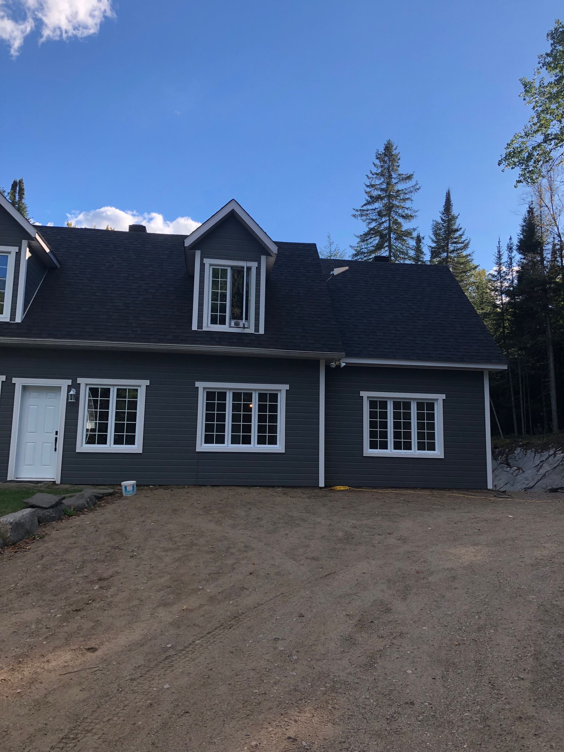 Une maison de deux étages gris anthracite, avec des encadrements de fenêtres blancs et une lucarne, se détachant sur un fond d'arbres et de ciel bleu.