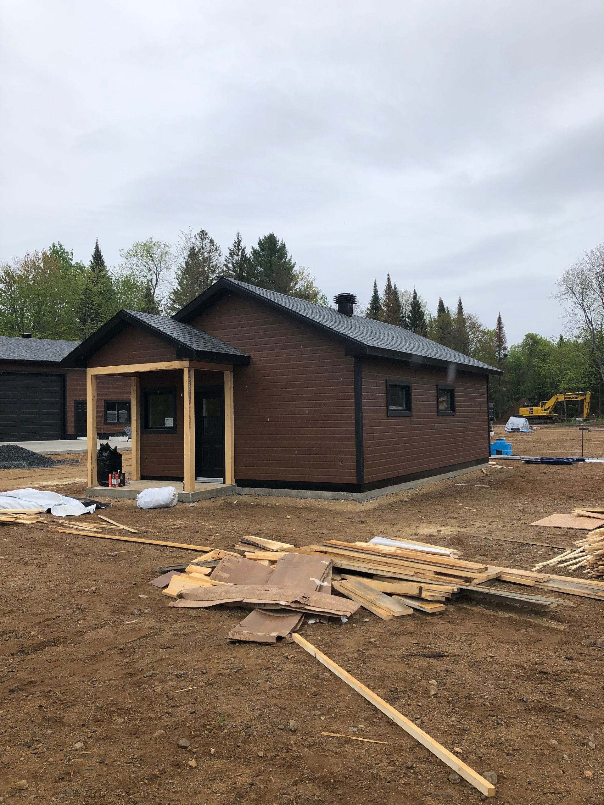 Une cabane brune au toit sombre et dotée d'une petite véranda couverte se dresse sur un chantier de construction jonché de débris et d'arbres.