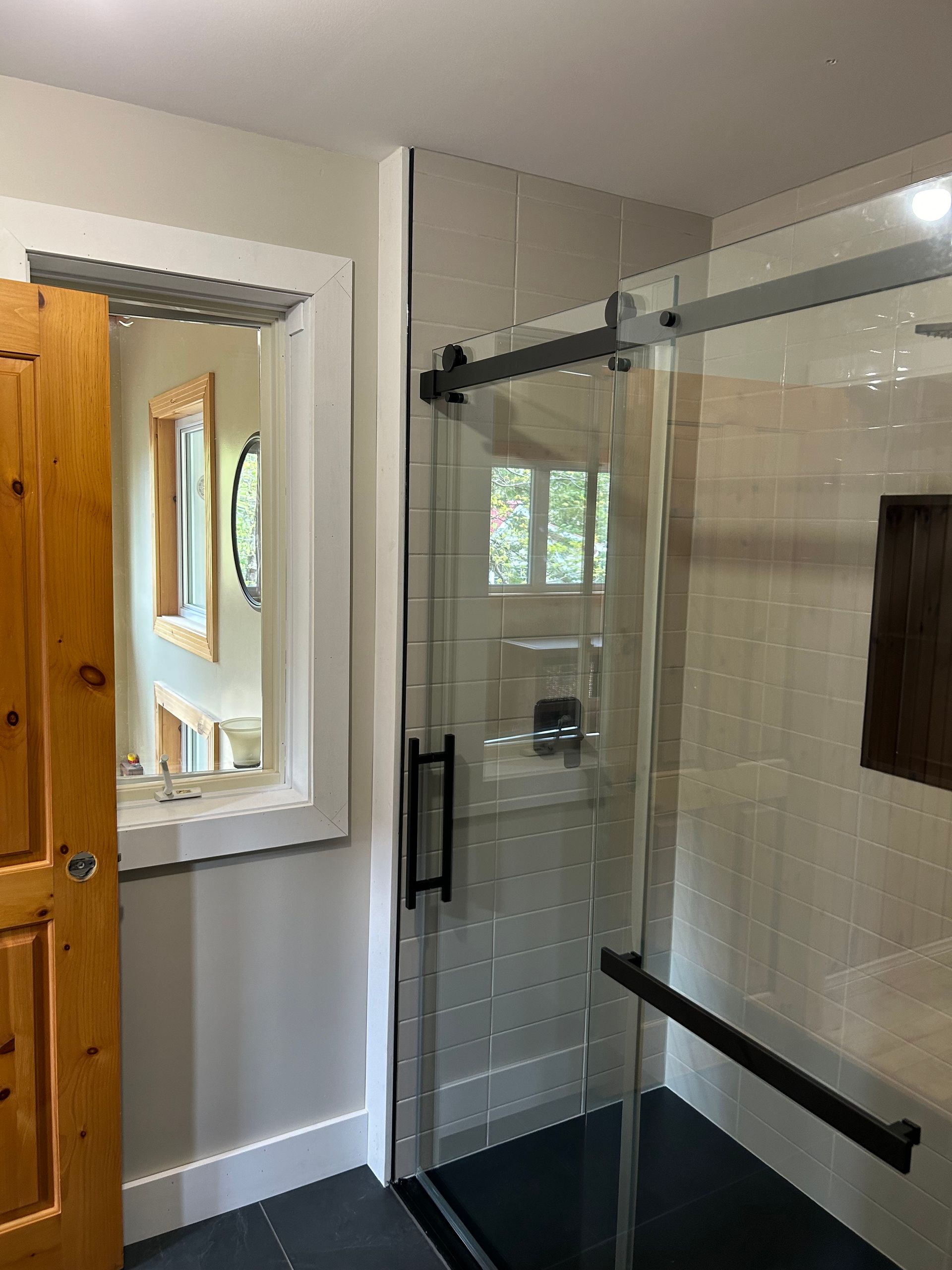 Une salle de bains moderne dotée d'une cabine de douche en verre avec robinetterie noire, à côté d'un miroir mural encastré reflétant une fenêtre.