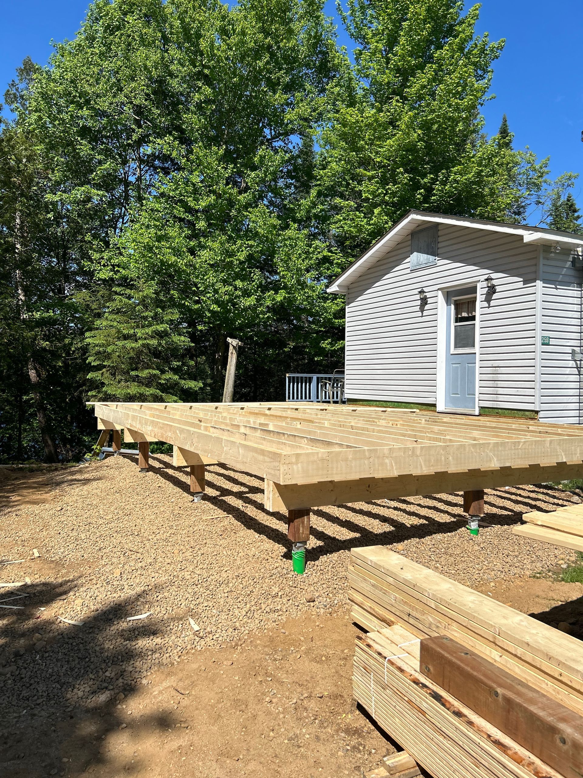 Une structure de terrasse en bois en construction sur du gravier, située devant une maison blanche entourée d'arbres par une journée ensoleillée.