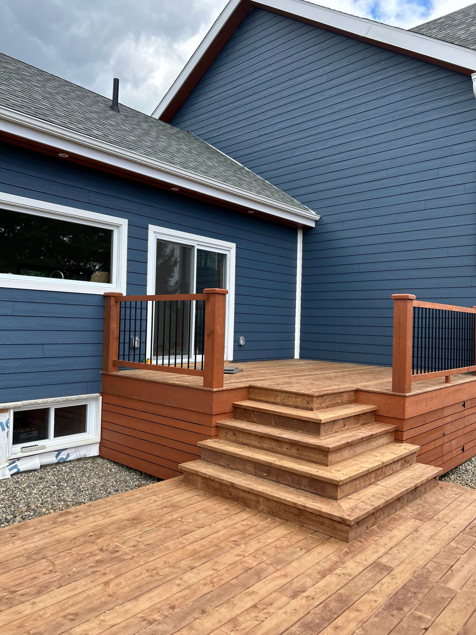 Une terrasse avec des escaliers et des rambardes couleur bois, adossée à une maison au bardage horizontal bleu foncé.