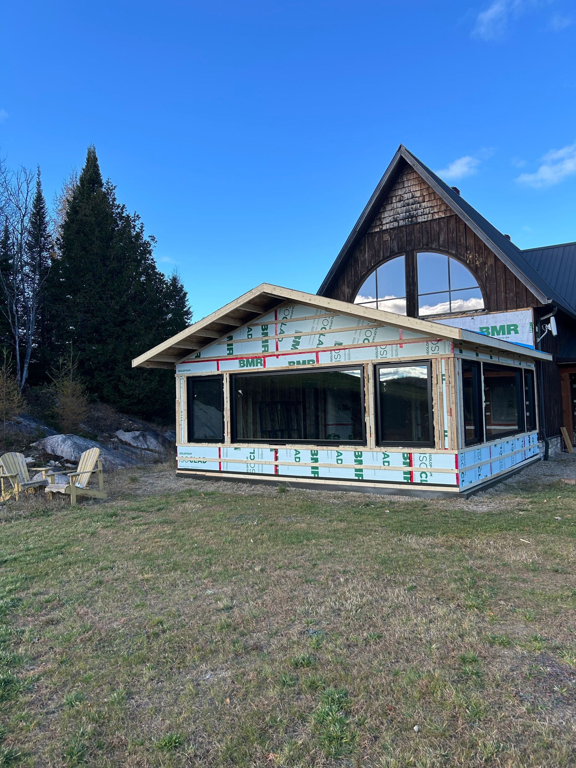 Une extension en bois partiellement achevée, avec des fenêtres déjà installées, est adossée à une maison plus grande sous un ciel d'un bleu éclatant.