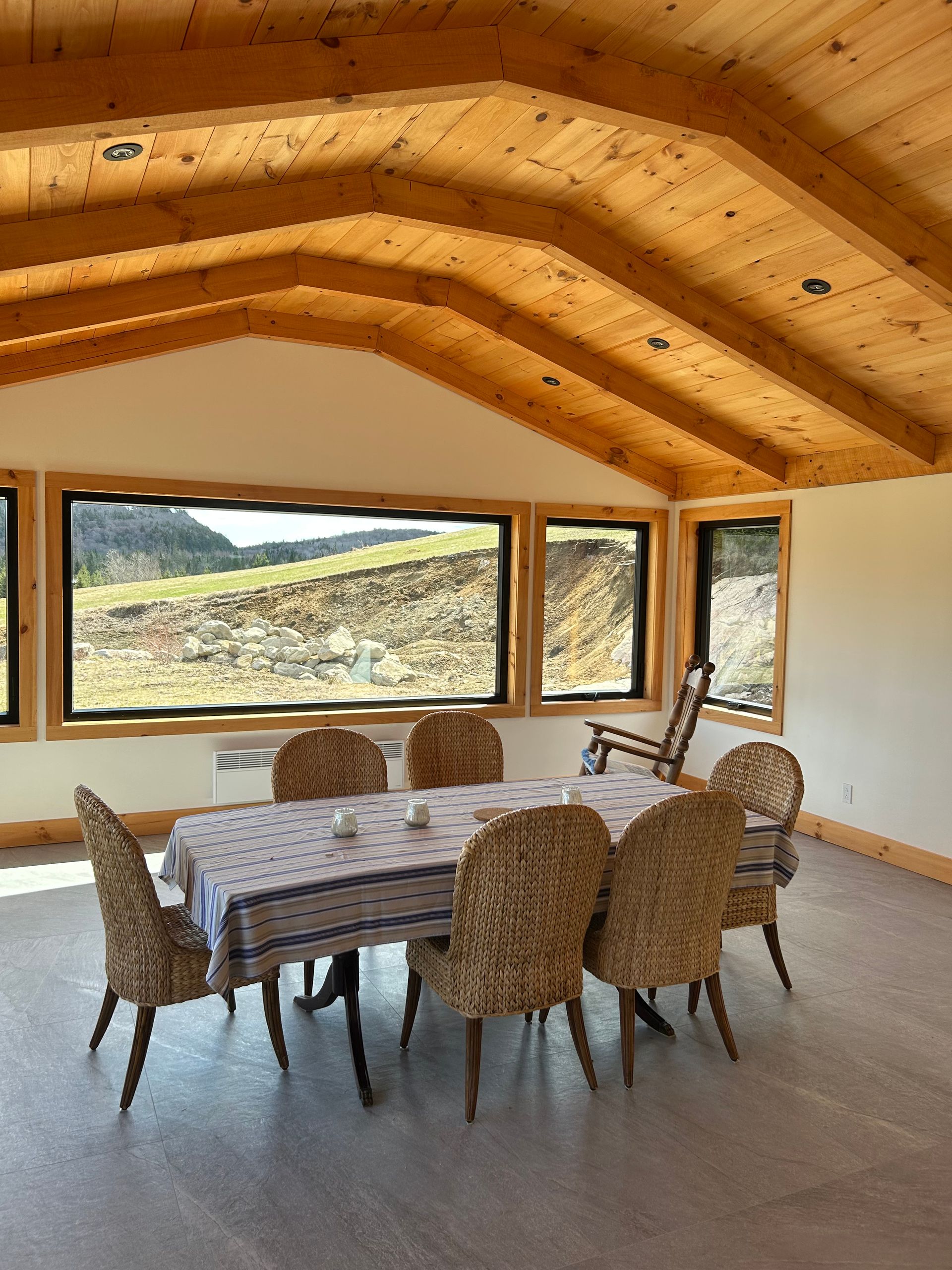 Salle à manger avec table, chaises en osier et plafonds voûtés en bois, donnant sur un paysage grâce à de grandes fenêtres.
