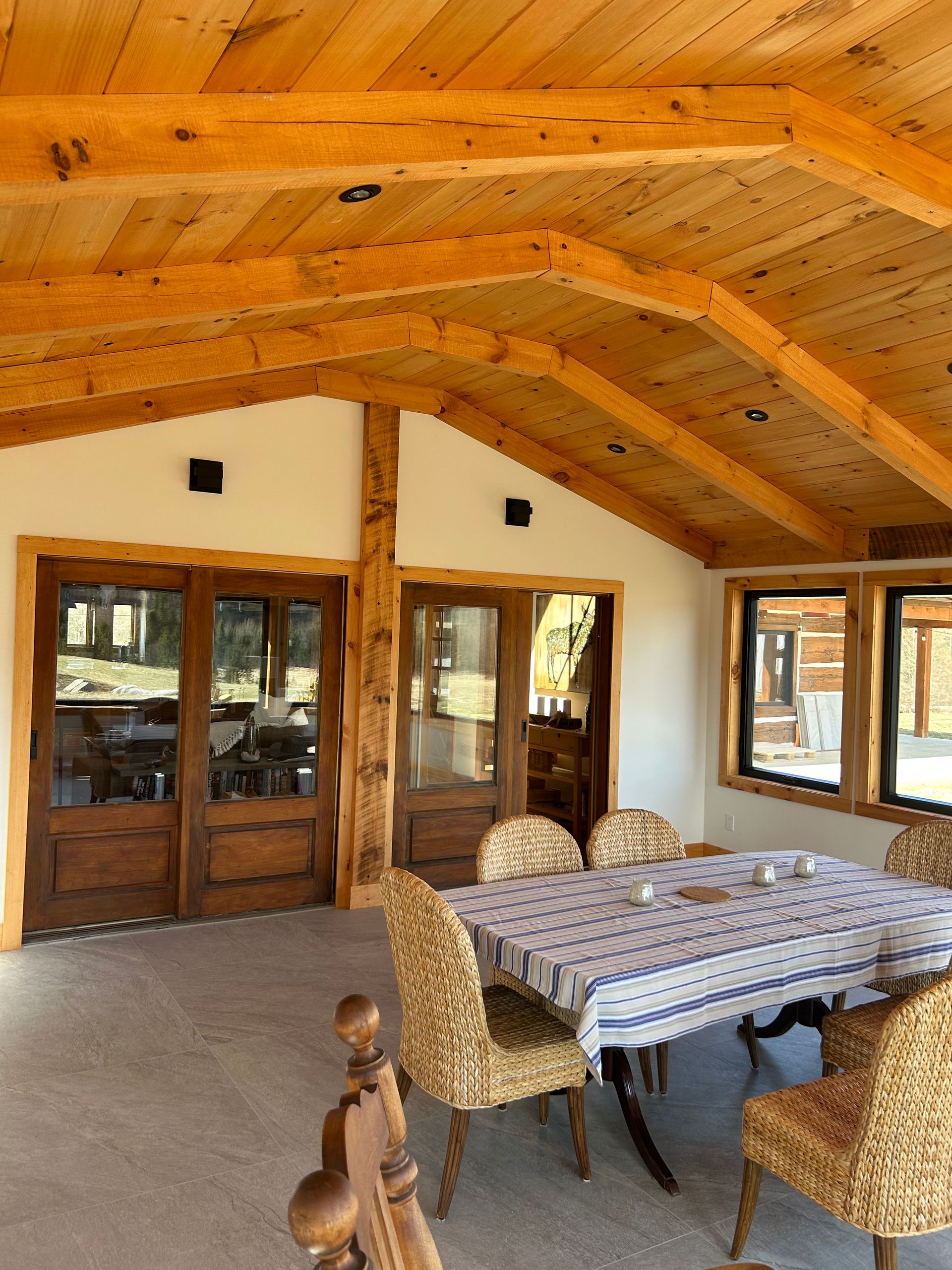 Coin repas sur une véranda grillagée avec plafond à poutres apparentes, nappe à motifs et chaises en osier.