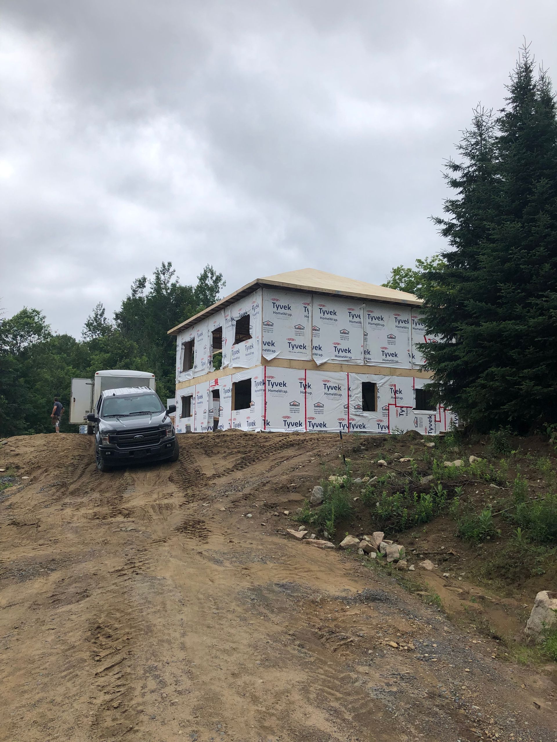 Un chantier de construction montrant la structure d'une maison à deux étages recouverte d'une bâche de protection blanche, avec un camion garé sur un talus de terre.