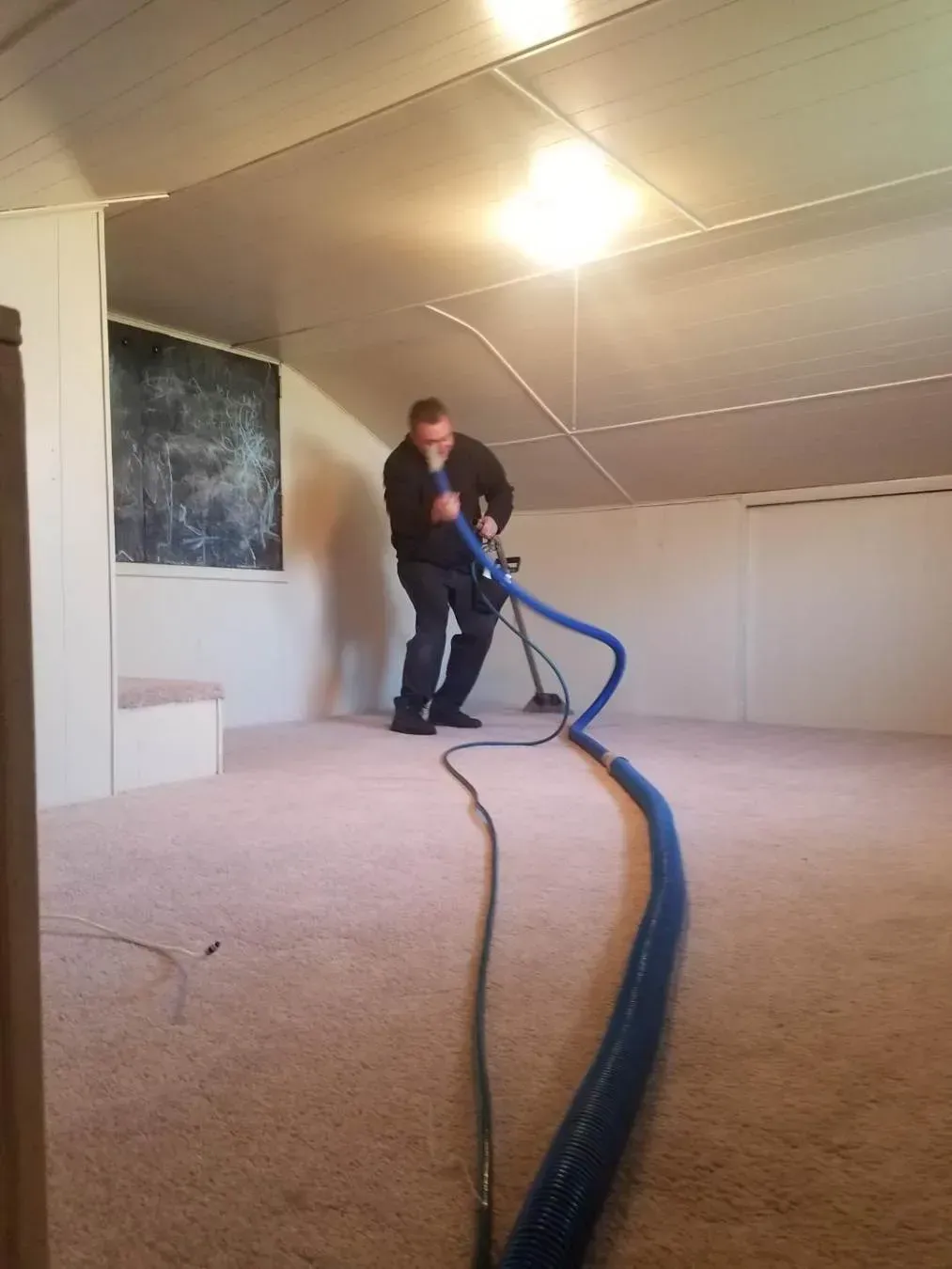 Man cleaning carpet with a machine in a room with beige carpet, white walls, and a chalkboard.