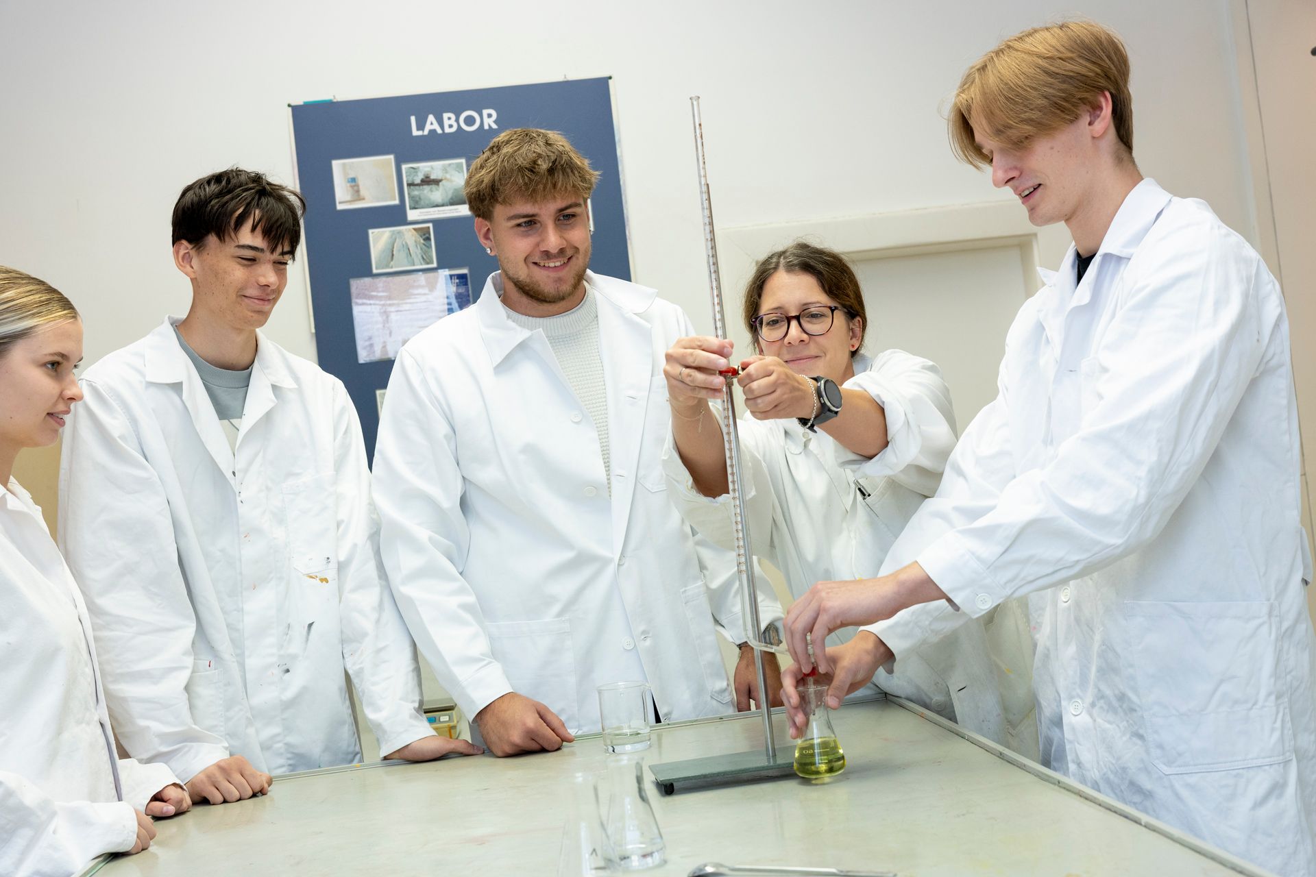 Fünf Studenten in weißen Laborkitteln arbeiten in einem hellen Klassenzimmer gemeinsam um einen Tisch mit Laborgeräten.
