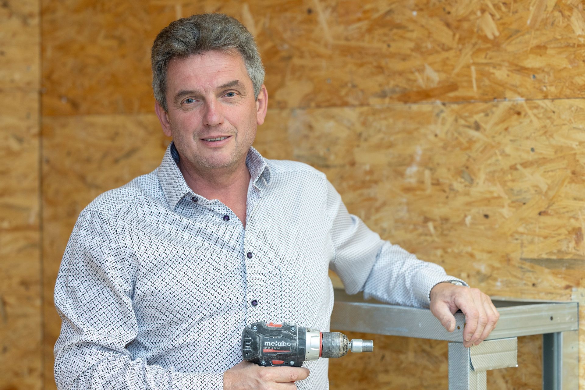 Person in einem gemusterten Button-Down-Hemd hält eine Bohrmaschine in der Hand und steht vor einer Wand aus OSB-Platten.