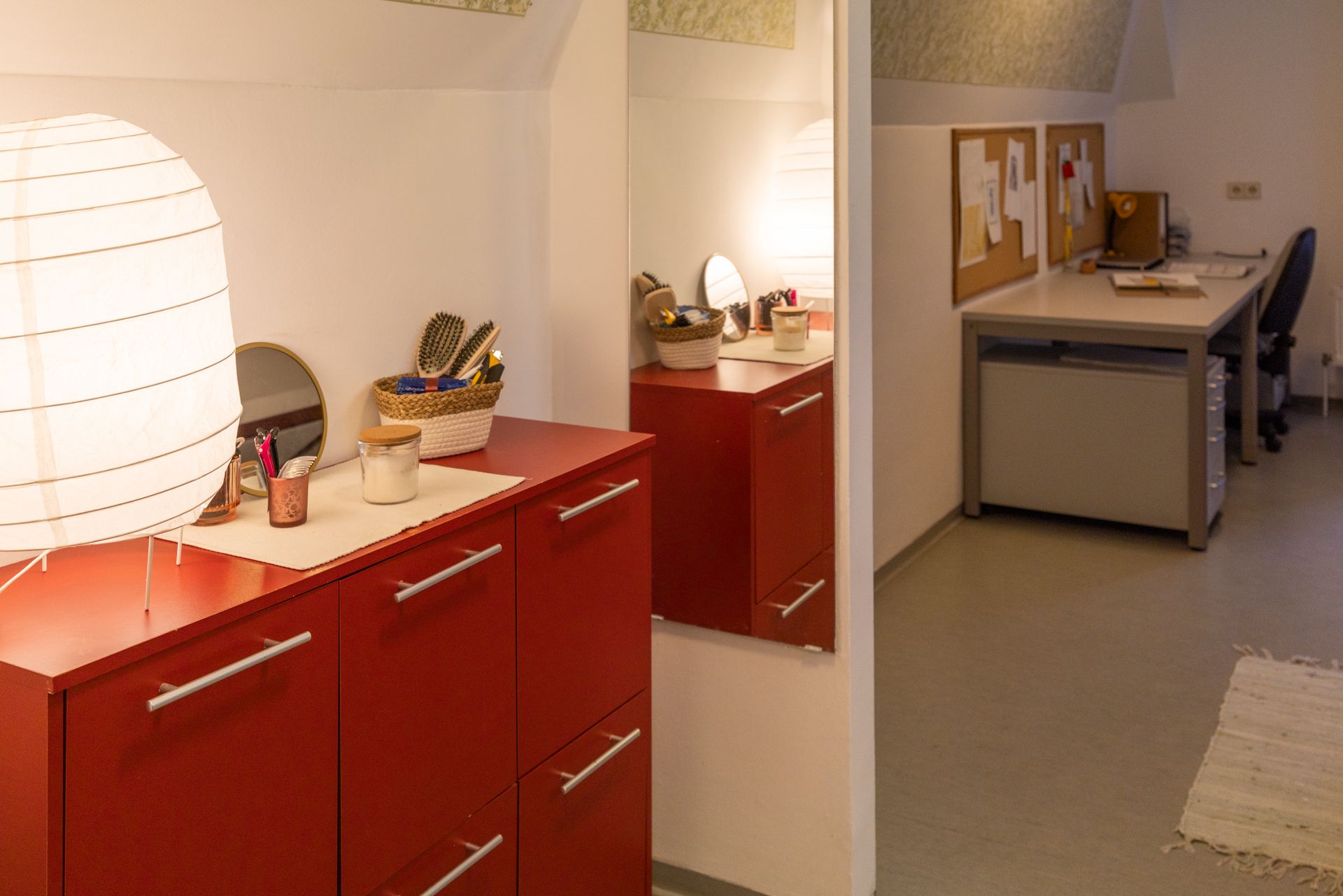 Im Vordergrund ein roter Schrank mit weißer Lampe, Spiegel und Toilettenartikeln, daneben Schreibtisch und Stuhl.