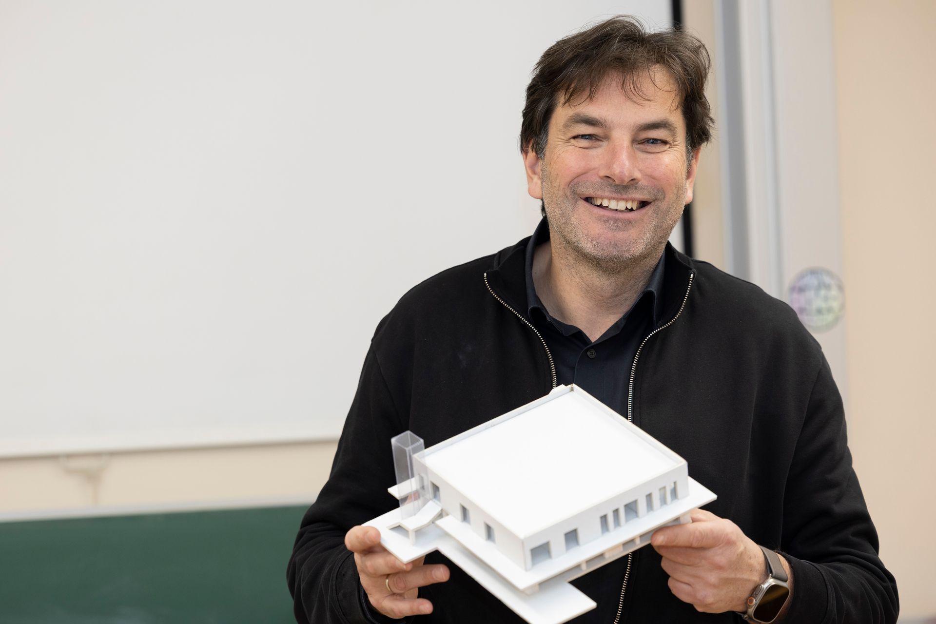 Eine lächelnde Person in einem schwarzen Pullover hält in einem Innenraum ein weißes Architekturmodell eines Hauses.