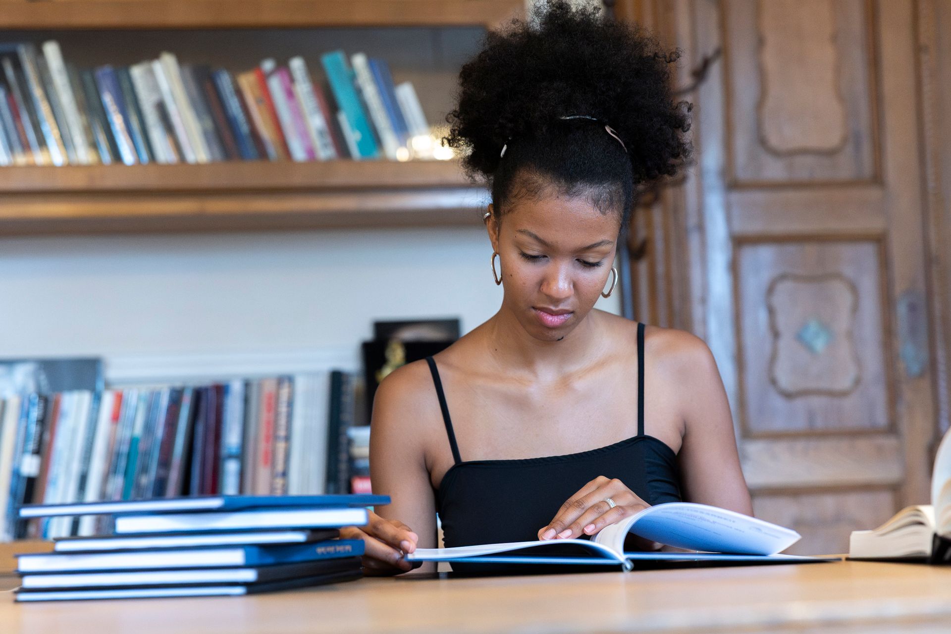 Person in schwarzen Oberteil sitzt an einem Schreibtisch und liest ein Buch; Hintergrund Bücherregale
