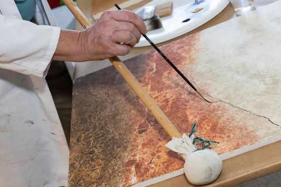 Eine Hand benutzt langen Malstock und einen Pinsel, um Farbe auf eine strukturierte, marmorierte Oberfläche aufzutragen.