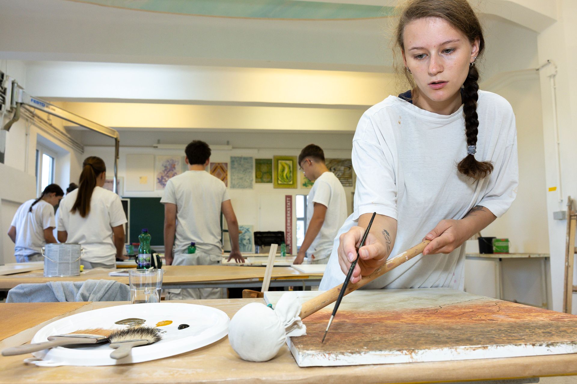 Die Schüler arbeiten in einem Kunstatelier; im Vordergrund malt eine Person mit einem Malstock auf einer Leinwand.