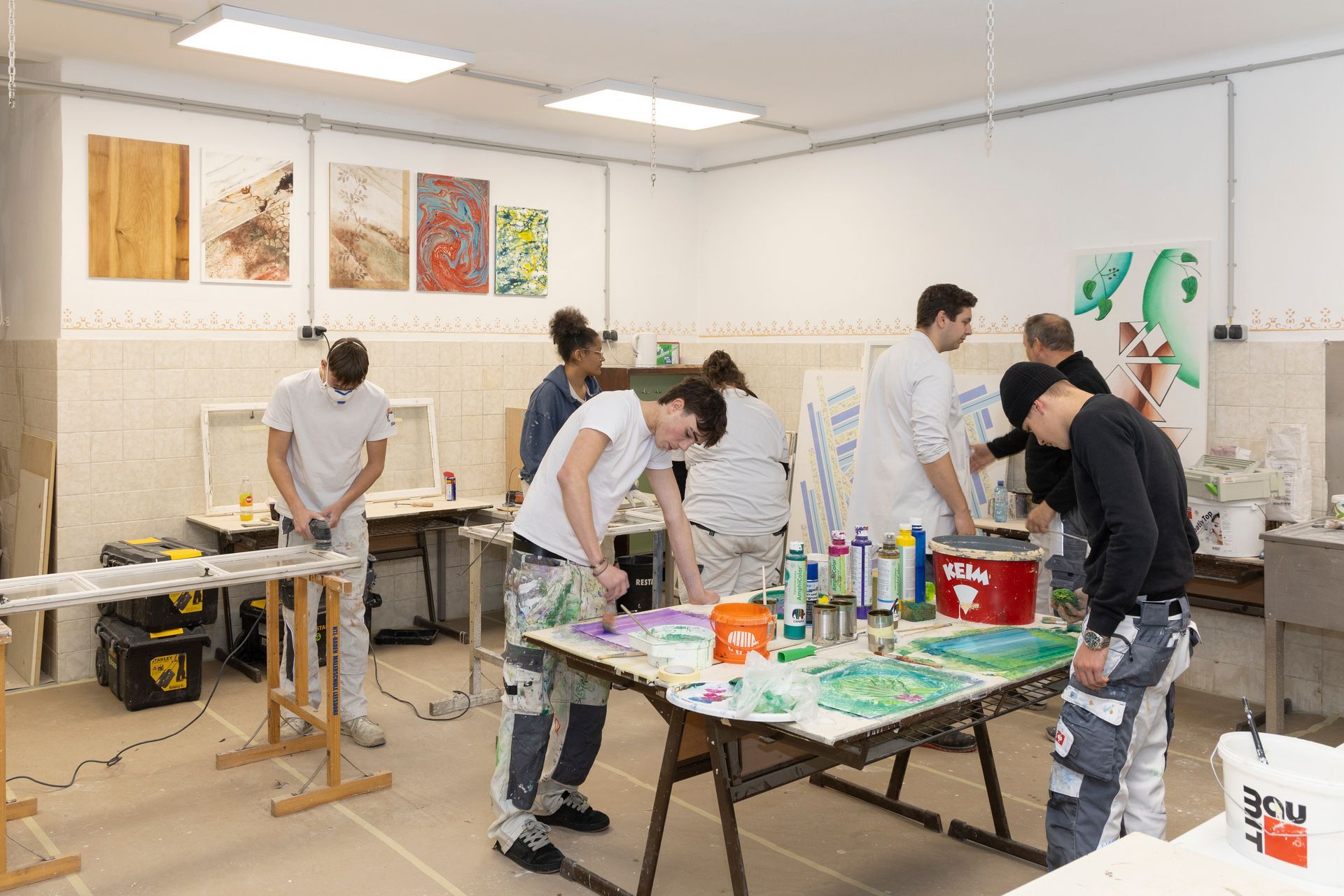 Studenten in Arbeitskleidung üben Maltechniken an Tischen in einem hell erleuchteten Atelier, an Wänden Kunstwerke hängen.