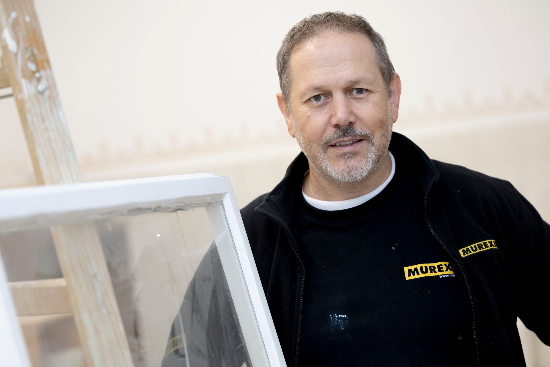 Ein Mann mit kurzem braunem Haar und Spitzbart, der eine schwarze Murex-Jacke trägt, steht in einem Innenraum