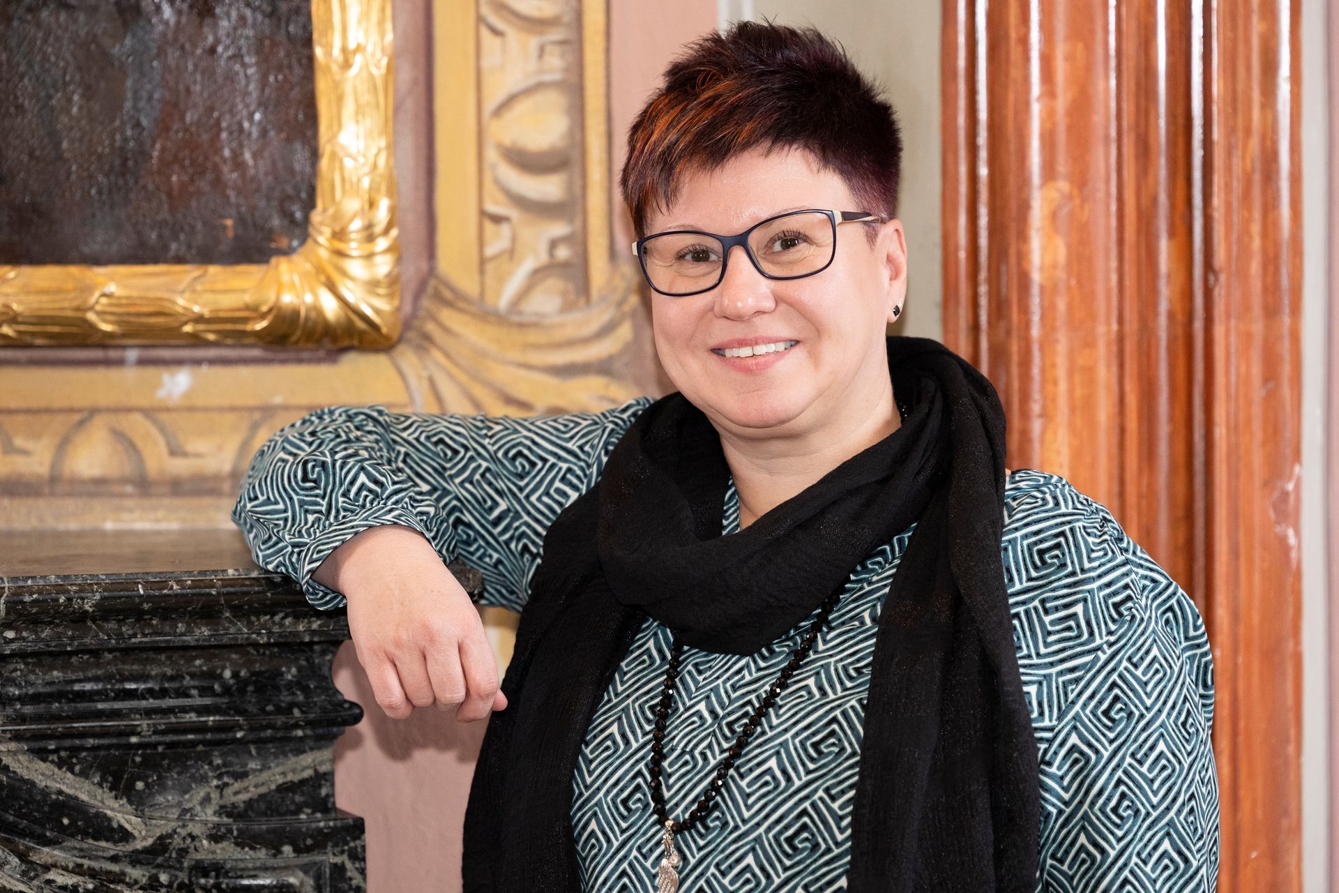 Eine Person mit kurzem, dunkel gesträhntem Haar lächelt und trägt ein gemustertes Oberteil und einen schwarzen Schal.
