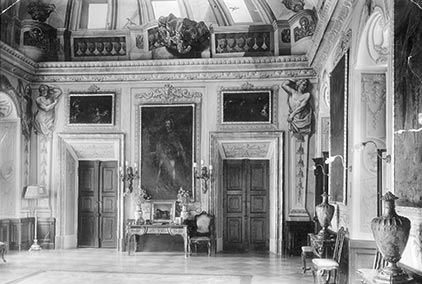 beigefarbenes, dreistöckiges historisches Herrenhaus mit Bogenfenstern, Steintreppe und Statuen in sonnigen Gartenanlage.