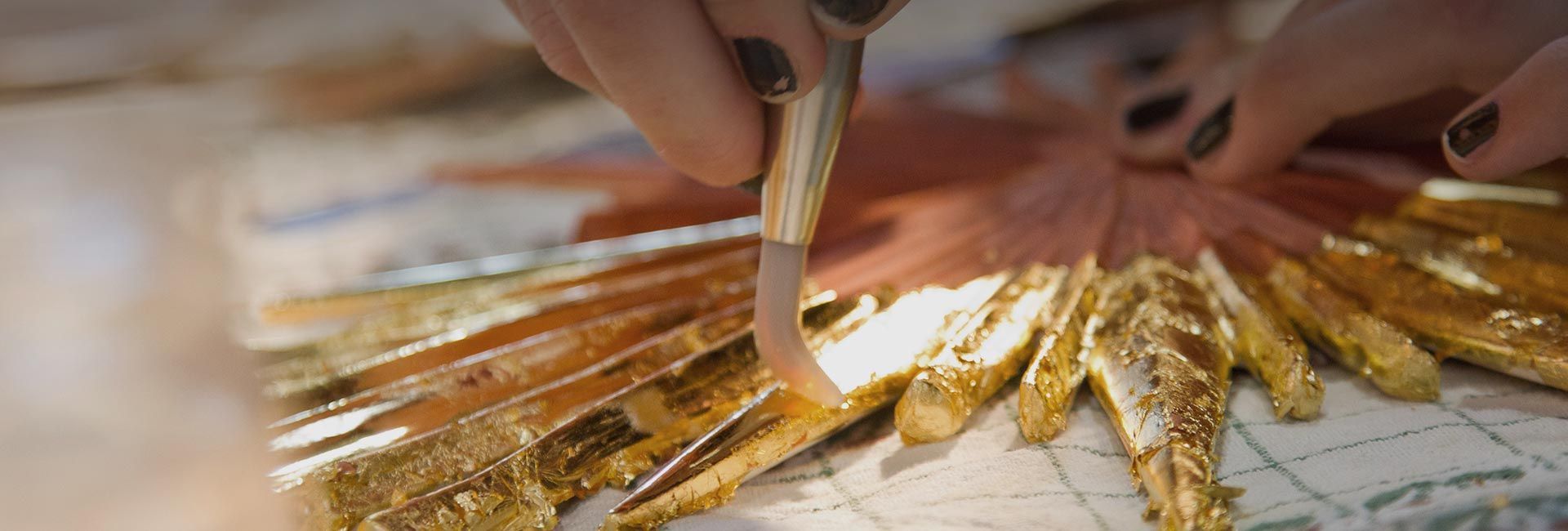 Mit einem Spezialwerkzeug tragen Hände sorgfältig Blattgold auf eine strukturierte, fächerförmige Oberfläche auf.