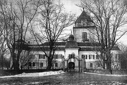 Schwarz-Weiß-Fotografie eines historischen Herrenhauses; zentraler Turm hinter kahlen Bäumen und eisernen Tor
