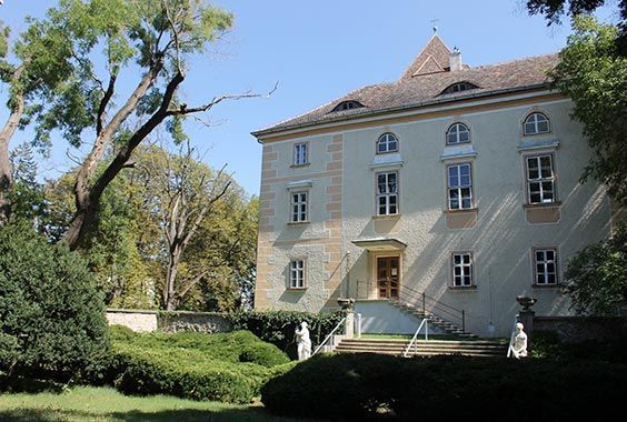beigefarbenes, dreistöckiges historisches Herrenhaus mit Bogenfenstern, Steintreppe und Statuen in sonnigen Gartenanlage.