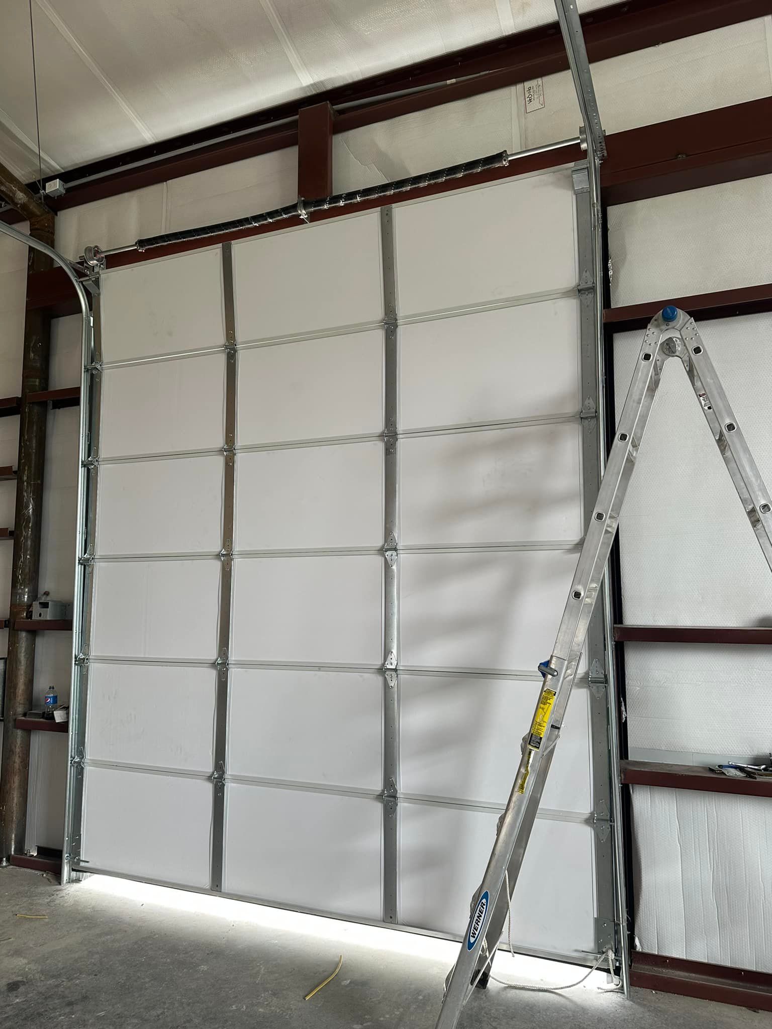 A large, white sectional garage door inside a workshop with metal wall framing and a ladder leaning against the right side.