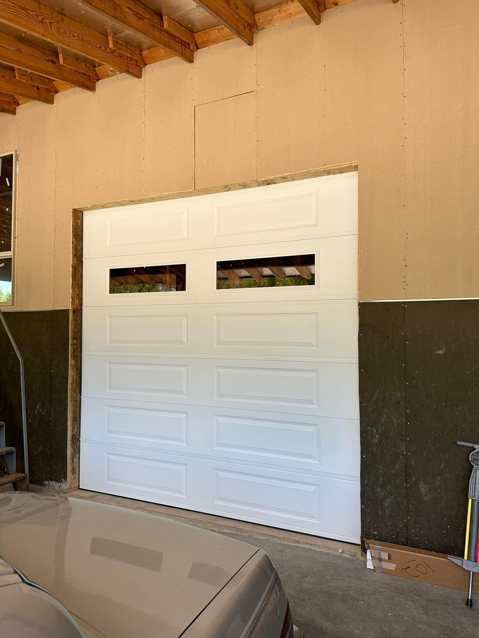 A white garage door with rectangular windows installed in a garage with unfinished wooden wall panels and exposed beams.