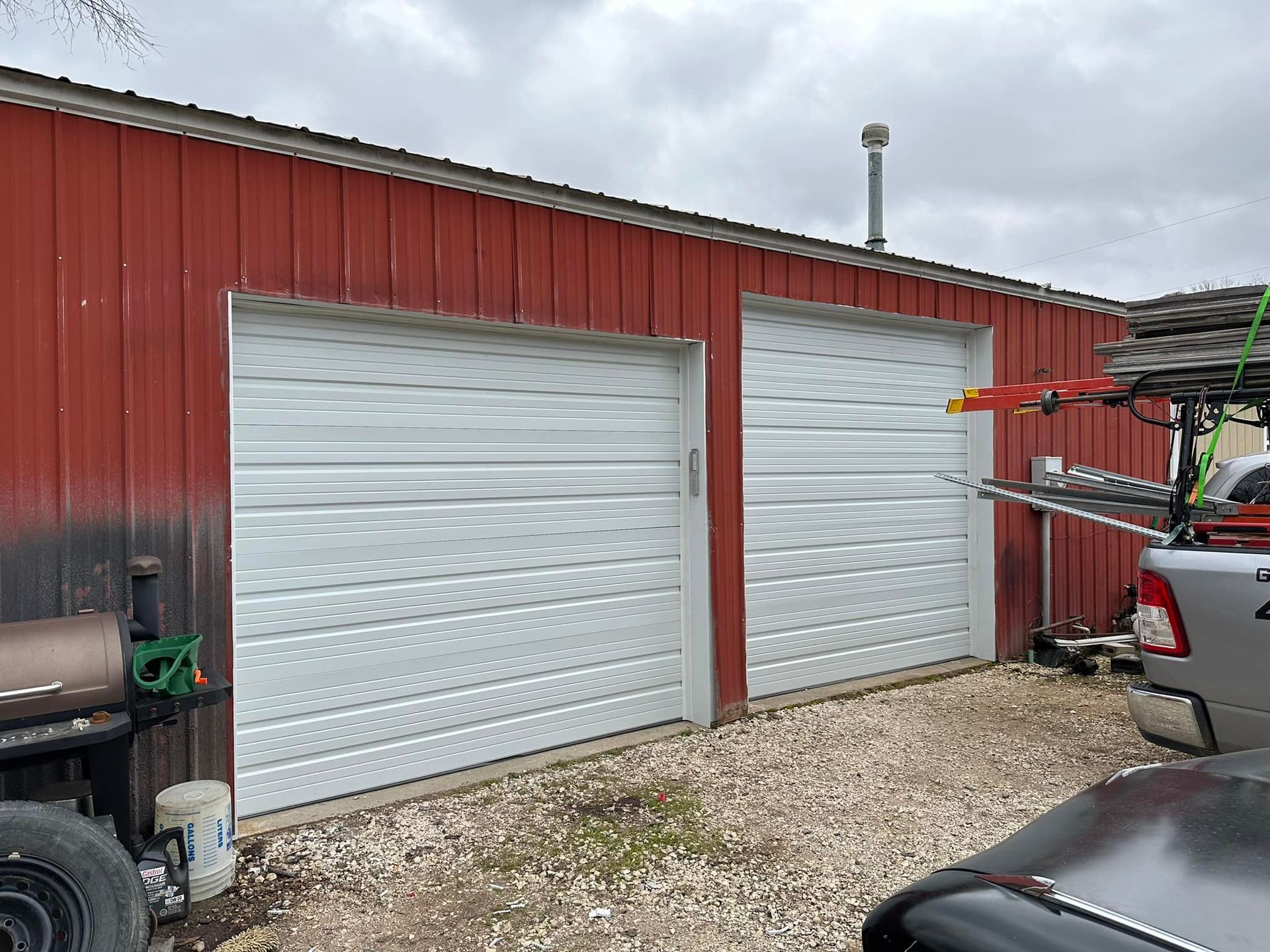 A red metal shed with two white roll-up garage doors, situated on a gravel lot near parked vehicles.