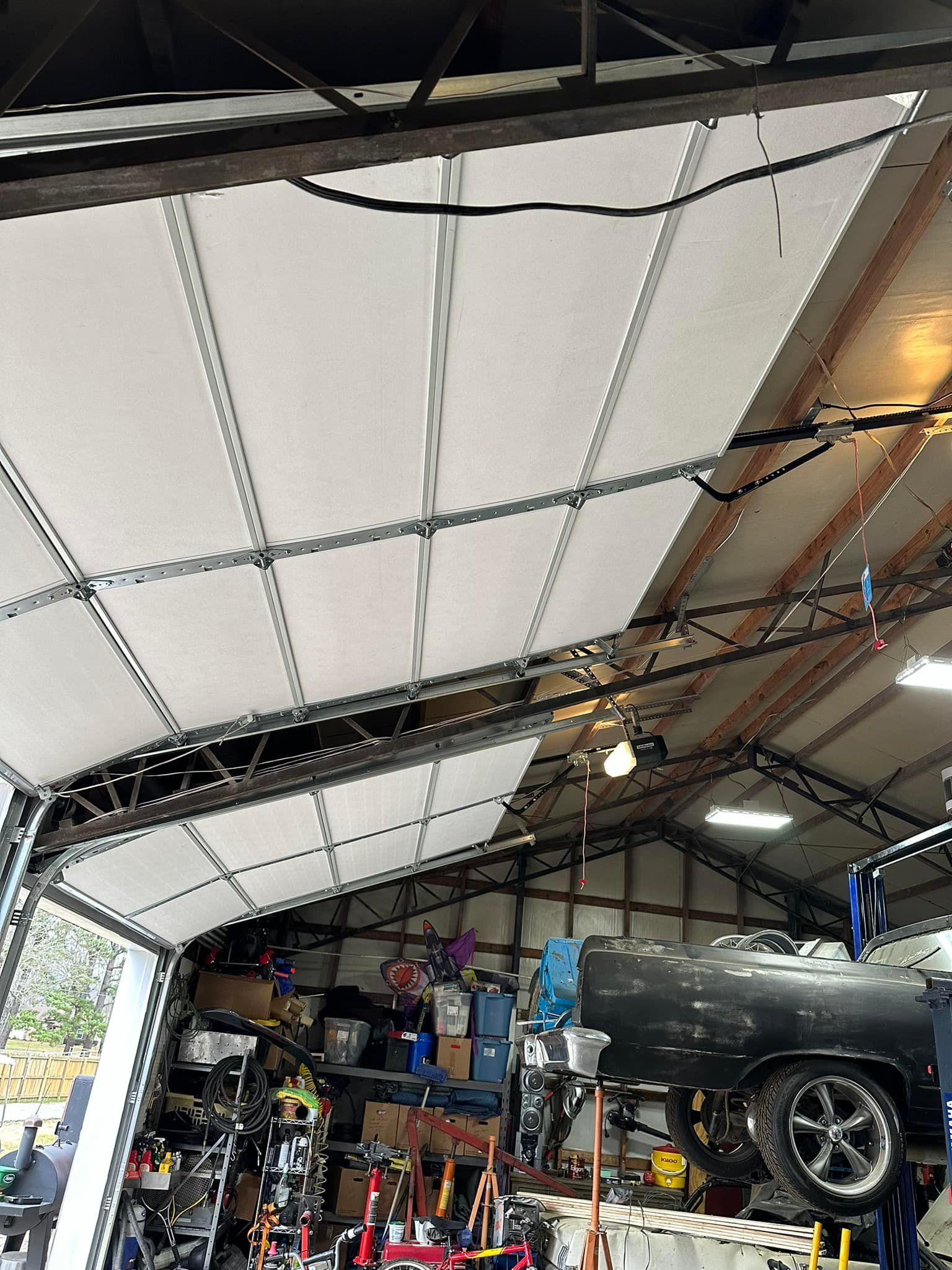 A white garage door open overhead in a cluttered workshop containing a car on a lift.