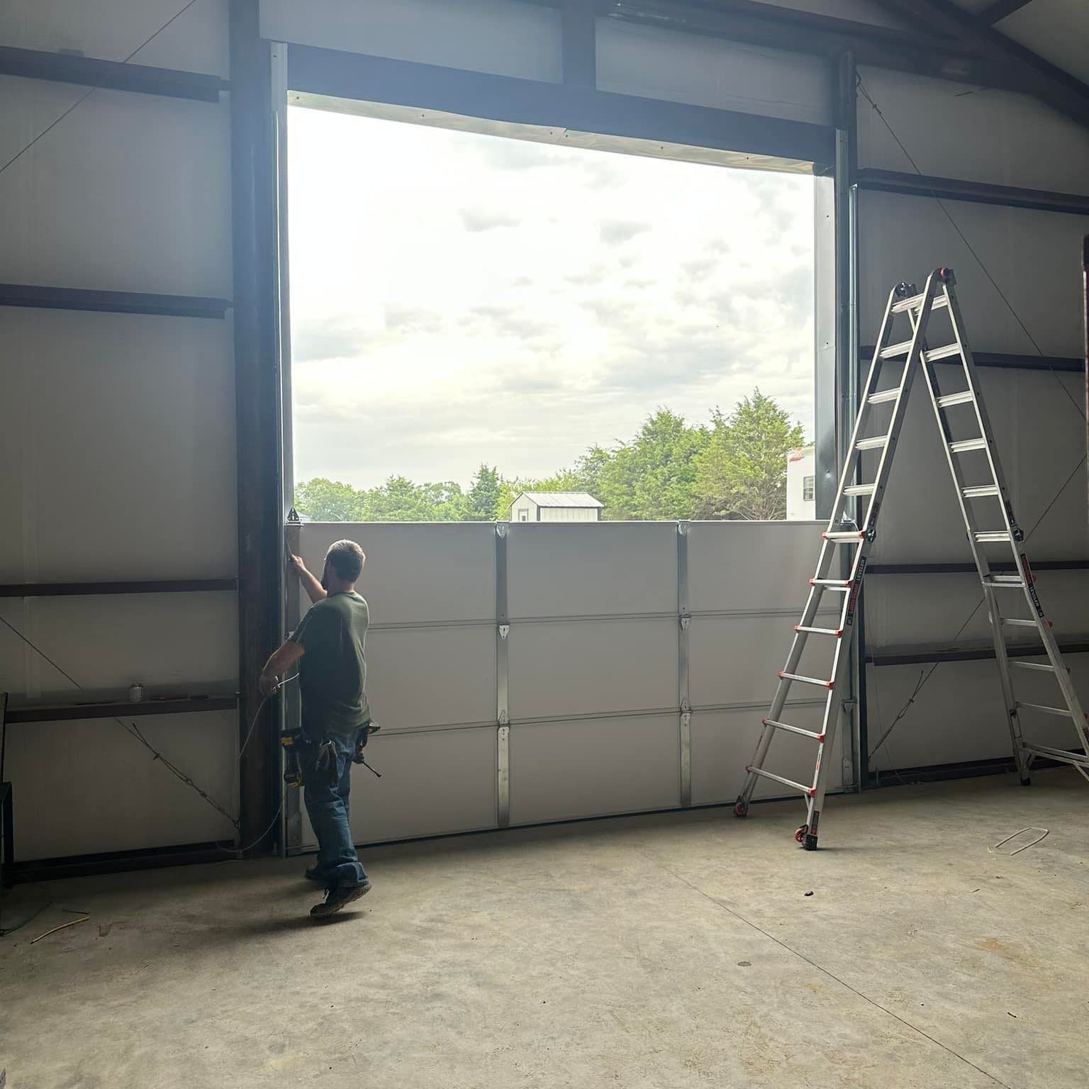 A person in work clothes stands inside a metal warehouse, adjusting large white panels near an open, full-height doorway.