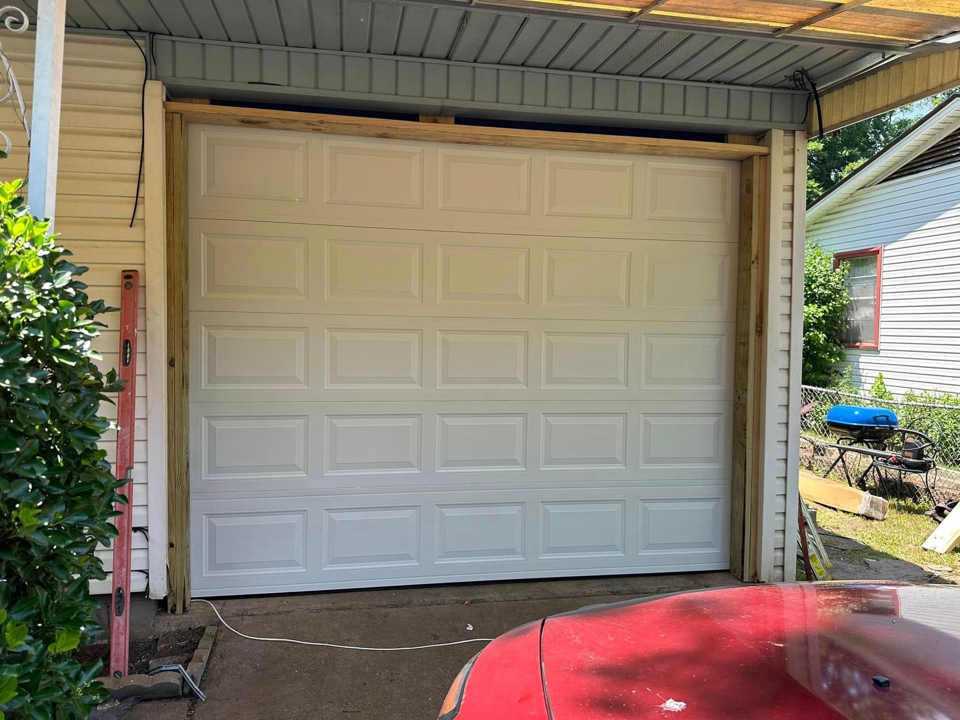 A white, multi-panel garage door installed in a wooden frame on the side of a building, with a red car hood in the foreground.