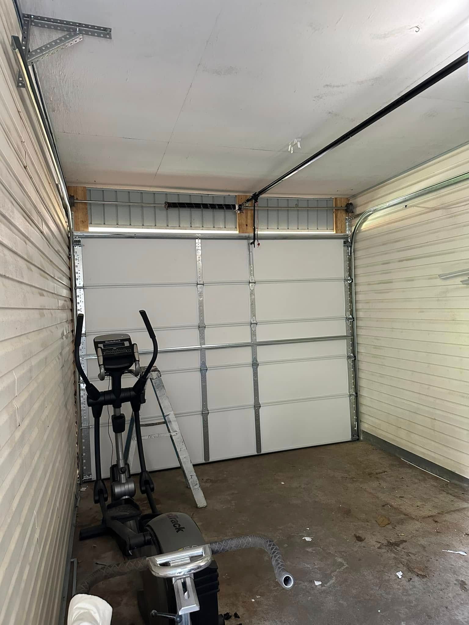 An elliptical machine sits inside a detached garage with white paneled walls, a closed metal door, and a step ladder.