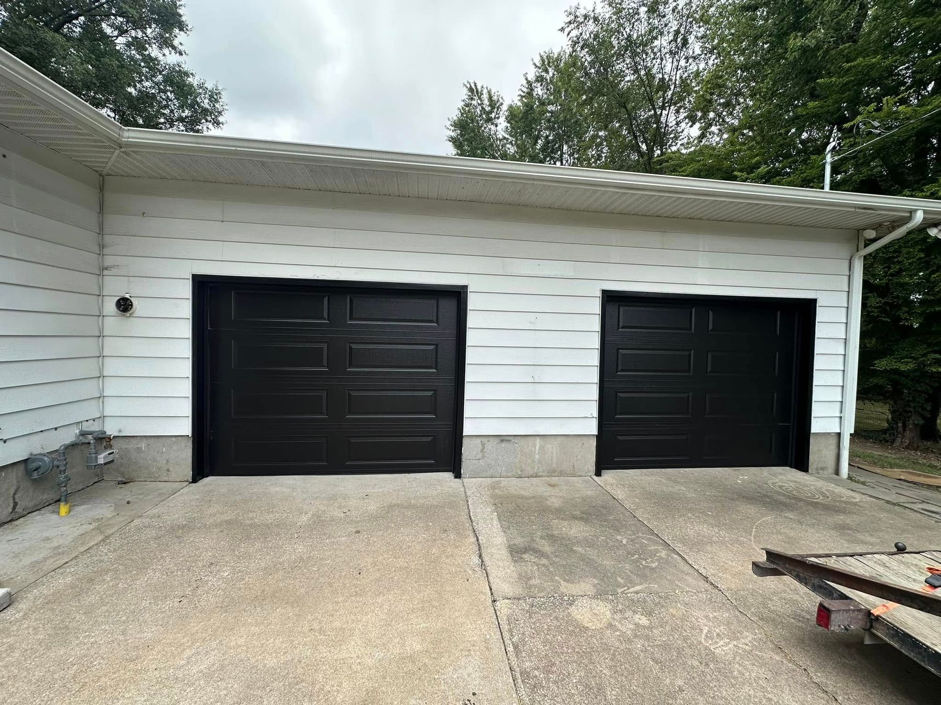 Two black garage doors installed in a white, horizontal-sided building with a concrete driveway in front.