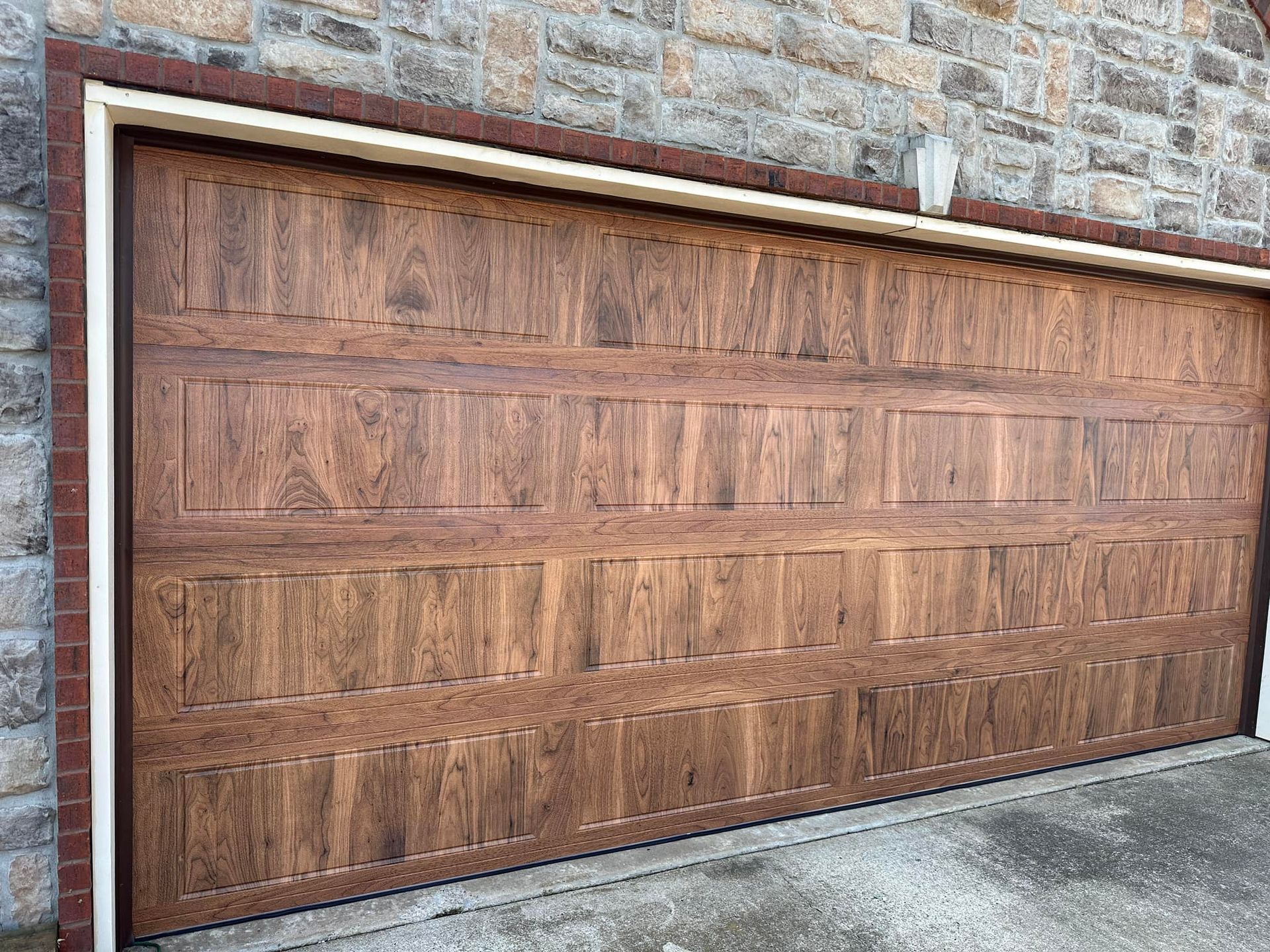 A brown, wood-grain patterned garage door with rectangular panel detailing, installed in a stone wall.