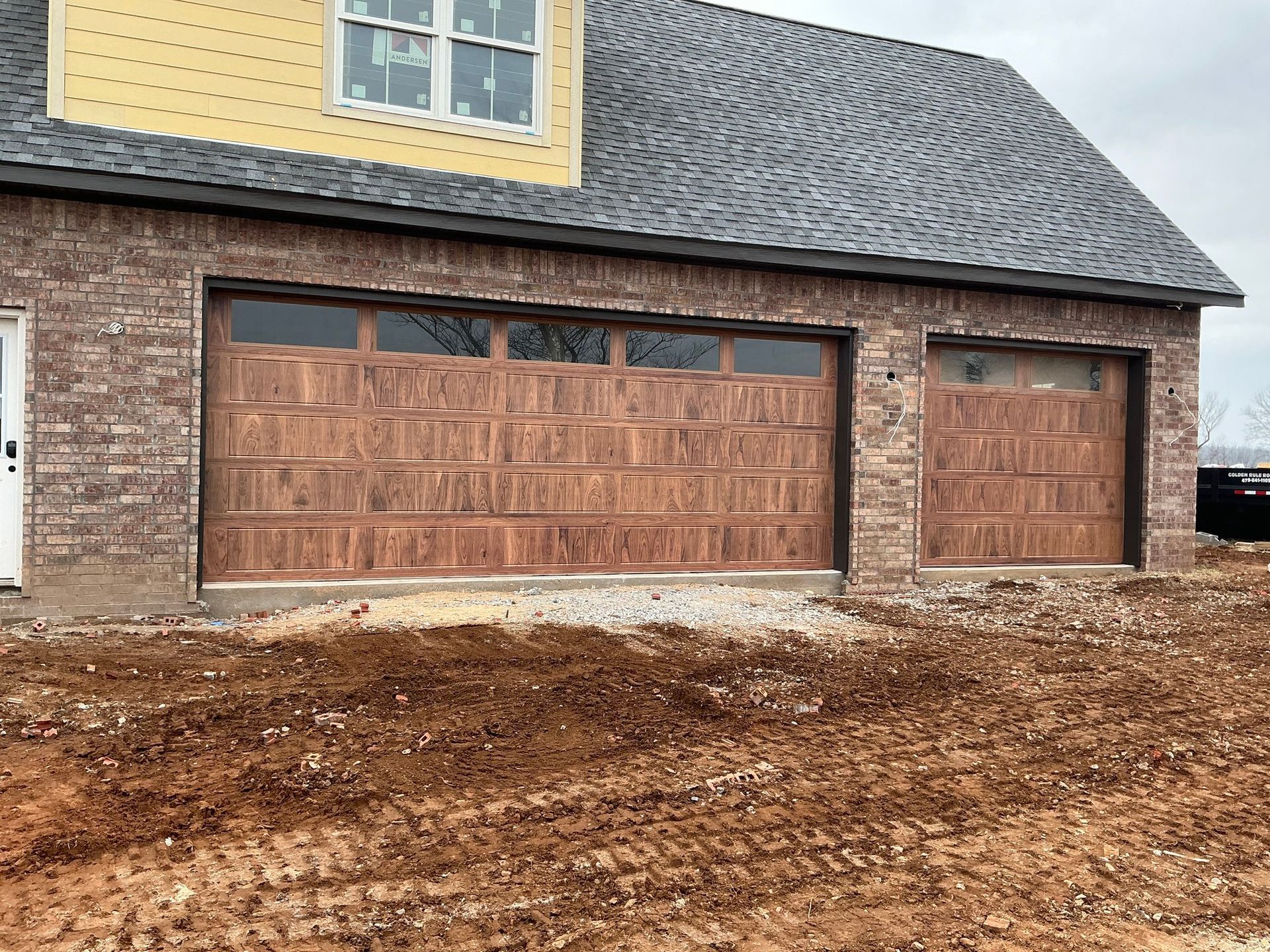 New house exterior with tan brick, a shingled roof, two wood-finish garage doors, and unfinished ground in front.