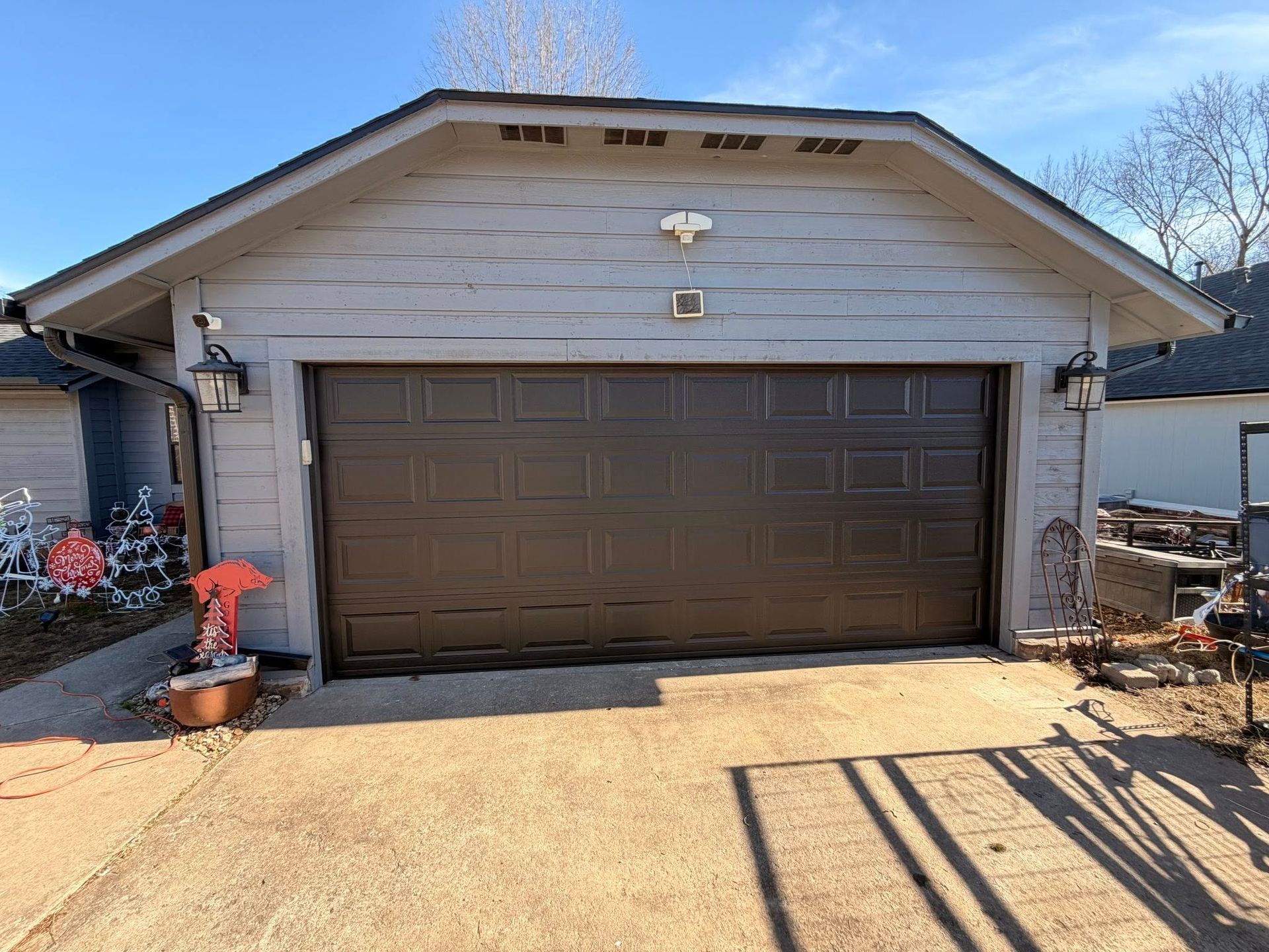 A detached gray garage with a dark brown, multi-panel door, two wall-mounted lanterns, and a concrete driveway.