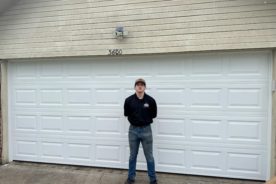 A person stands in front of a white garage door with rectangular paneling beneath a brick exterior wall.