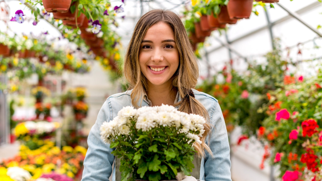 Woman smiling in a greenhouse, holding a pot of white flowers.