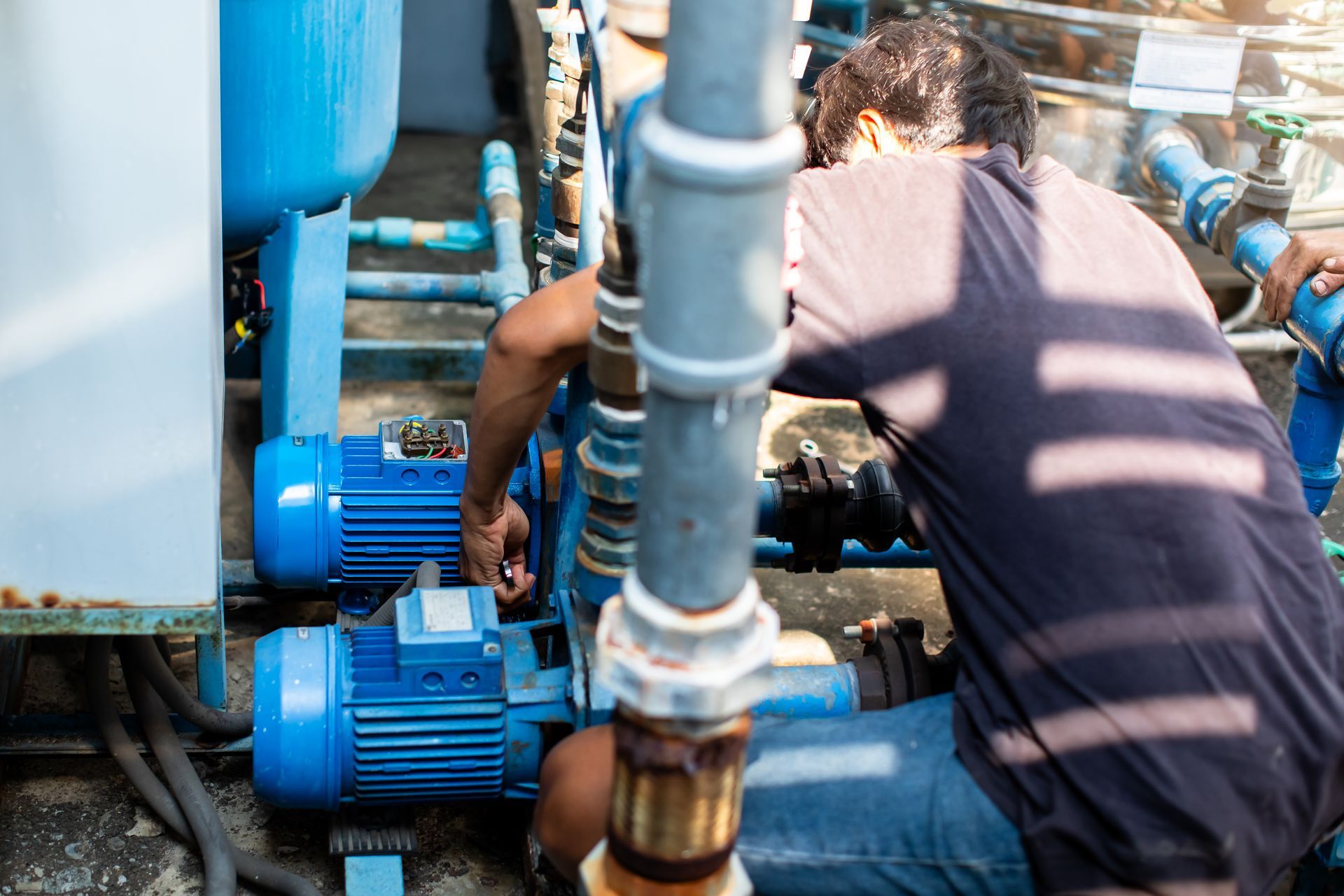 A man repairs a pump outdoors, surrounded by machinery and tools.