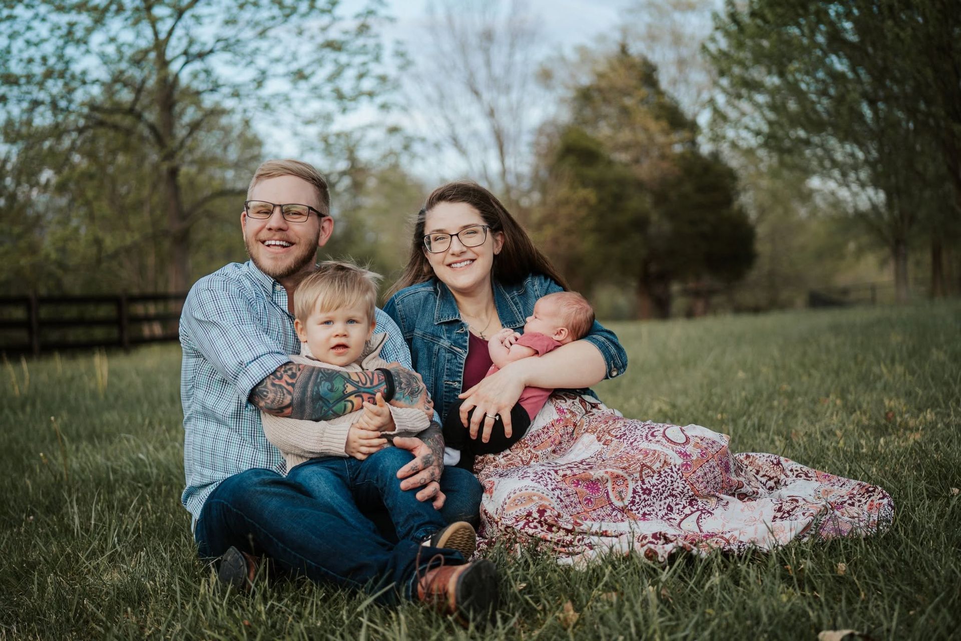 Family of four sitting on grass in a park, smiling and cuddling outdoors.