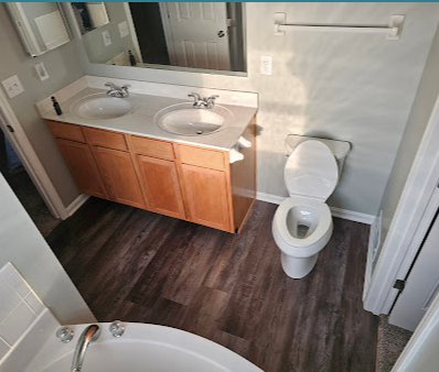 Small bathroom with double sink vanity, mirror, toilet, and visible bathtub corner with dark wood floor.