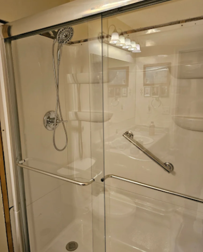 Clear glass walk-in shower with chrome grab bars and handheld showerhead in a beige bathroom