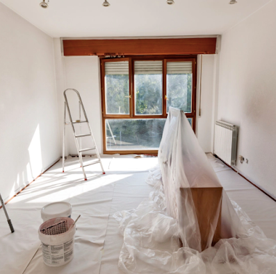 Sunlit room under renovation with plastic-covered furniture, ladder, paint bucket, and large window.