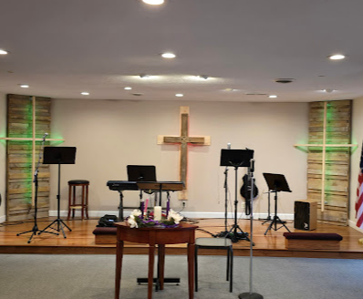 Church stage with cross, keyboard, microphones, music stands, and two stools under recessed lights