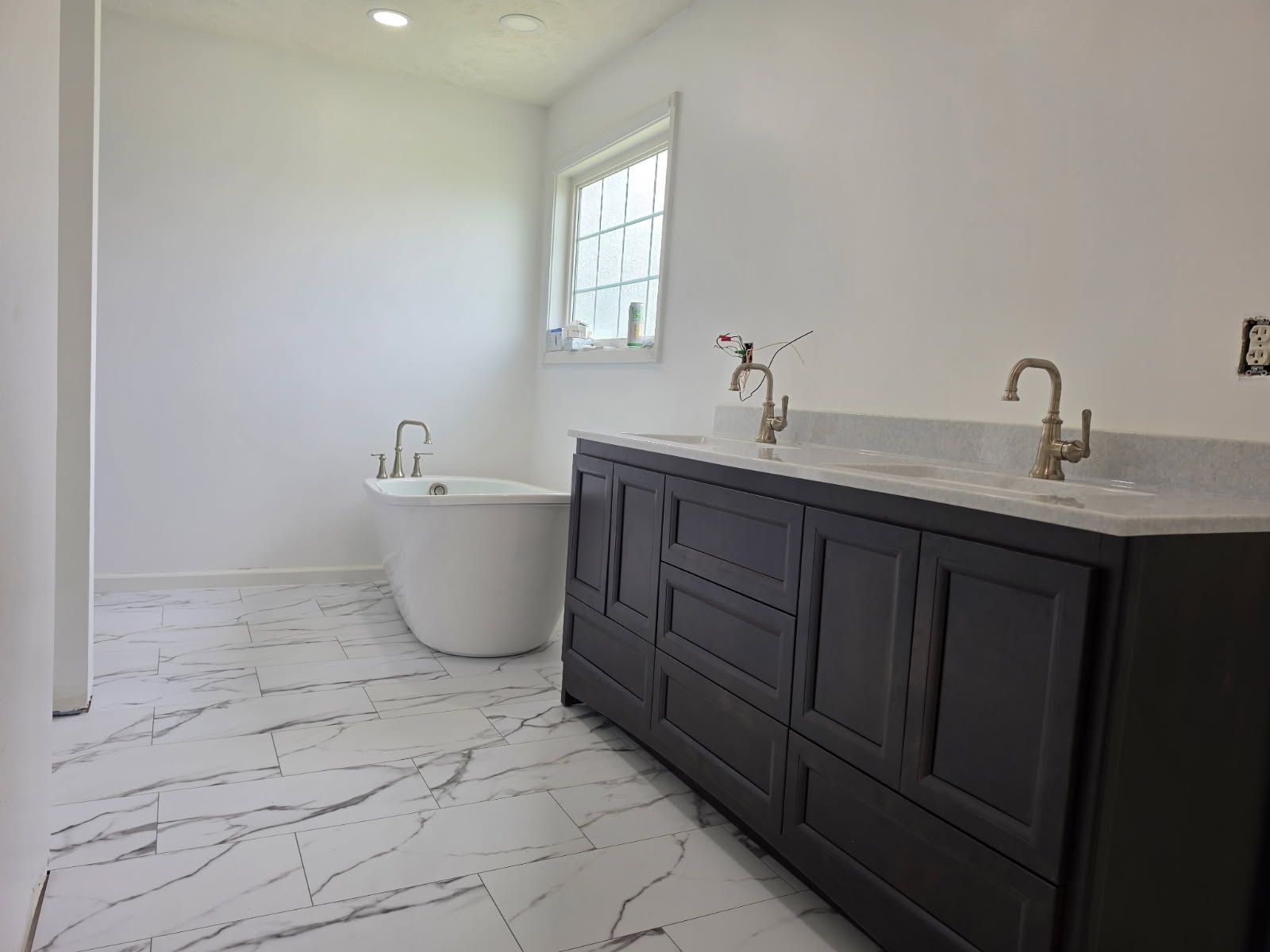 Bright modern bathroom with freestanding tub, dark vanity, brass faucets, and marble-look tile floor