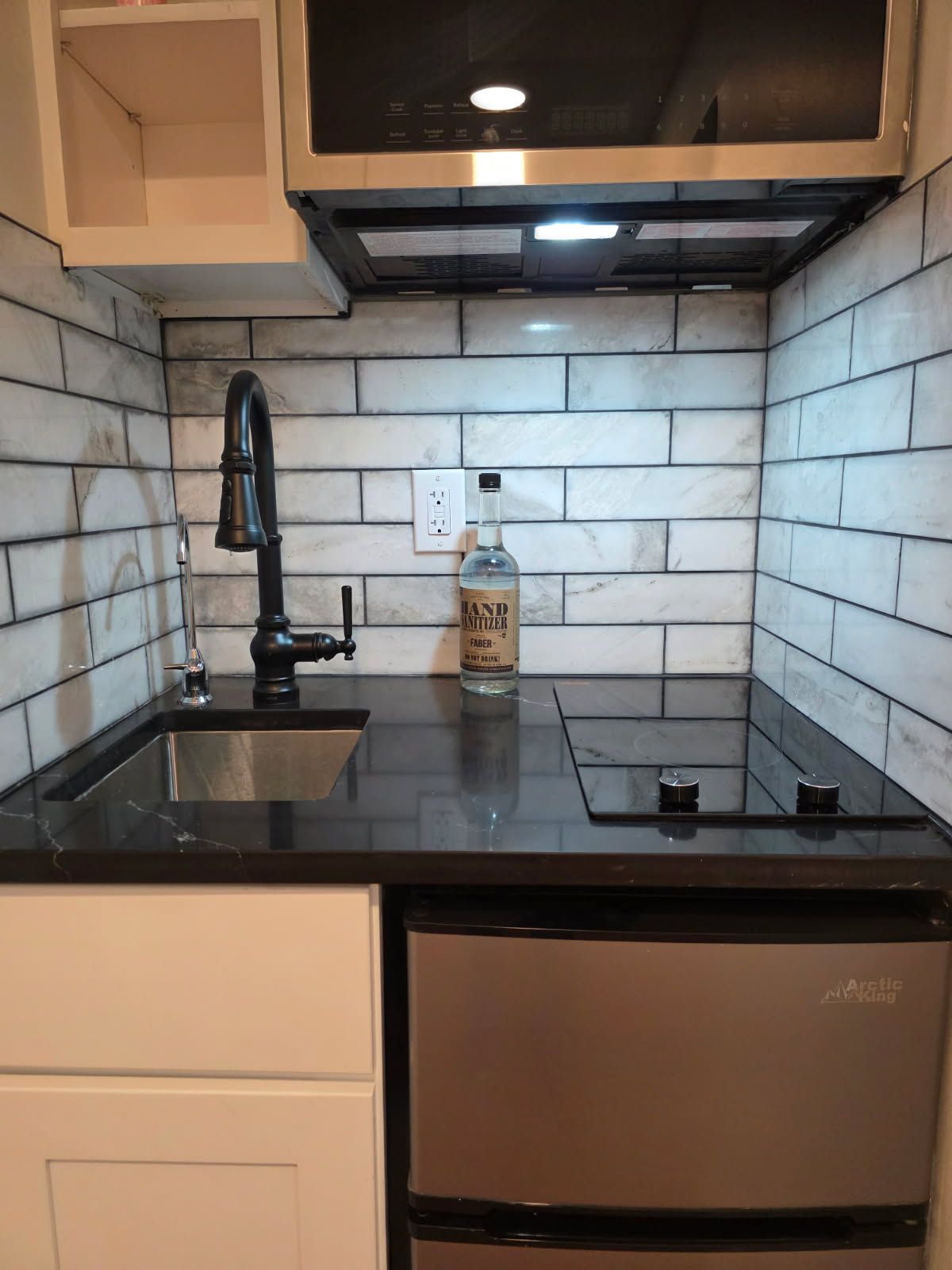 Small modern kitchen with black countertops, white tile backsplash, sink, stove, and stainless steel refrigerator.